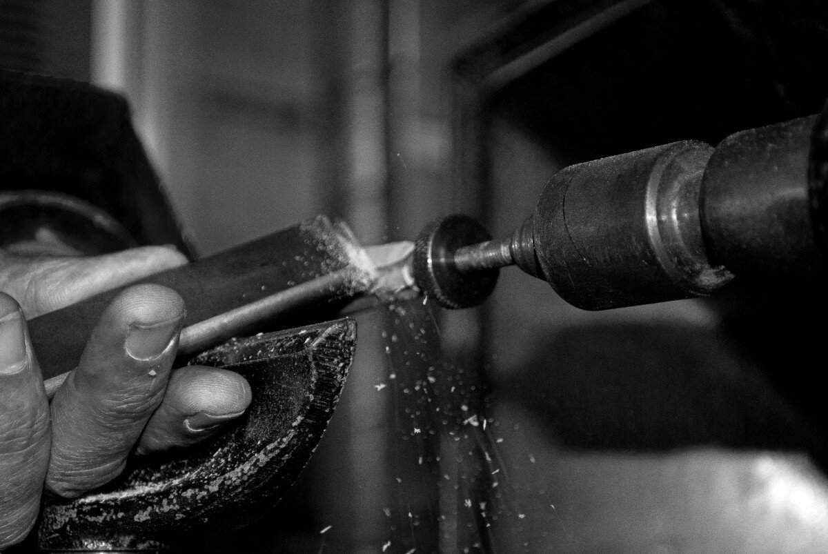 Fotografia czarno‑biała przedstawia wiertarkę z końcówką szlifującą, która szlifuje przedmiot trzymany przez rękę człowieka. Na szarym tle widoczne cztery palce człowieka. Palce trzymają ciemny podłużny metalowy przedmiot, który jednym końcem dotyka szlifierki. Widać sypiące się iskry.