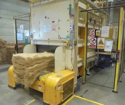 Zdjęcie przedstawia targarkę - maszynę do produkcji wyrobów z włókna kokosowego. Stoi w hali zakładu produkcyjnego. Po bokach jest zabezpieczona ogrodzeniem z siatki. Na siatce znaki ostrzegawcze. W przedniej części maszyny ułożone płaskie włókna kokosowe.
