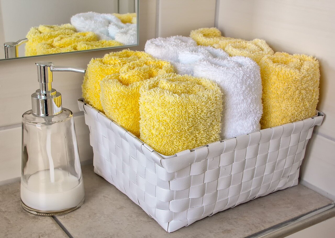 W łazience - od lewej mydło w płynie w dozowniku, kosz ze zwiniętymi w rulony ręcznikami, białe i żółte. 
