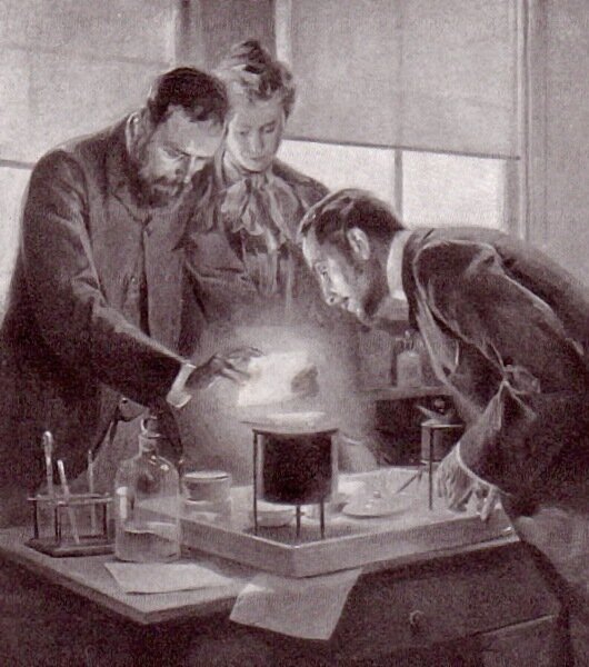 Ilustracja przedstawia trzy osoby, pochylające się nad stołem laboratoryjnym w czasie eksperymentu - dwóch mężczyzn i kobietę. Kobieta przygląda się z głębi ilustracji w centralnej części. Po prawej stronie mężczyzna pochyla się bliżej środka ilustracji. Z lewej strony mężczyzna w średnim wieku z brodą, podtrzymuje święcącą jasnym światłem płytkę. W tle widoczne przysłonięte okna. Na stole przyrządy laboratoryjne - pipety, butle. Widoczna szuflada pod stołem bliżej prawej krawędzi ilustracji. Ilustracja z epoki. 
