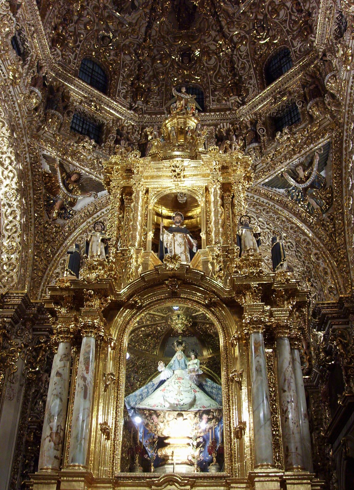 Ilustracja przedstawia barokowy ołtarz w meksykańskim kościele. Na ołtarzu stoi złoty tron Matki Bożej, który jest pokryty baldachimem. Głowice i podstawy kolumn są złote, podobnie jak łuki, pilastry oraz płaskorzeźby. 