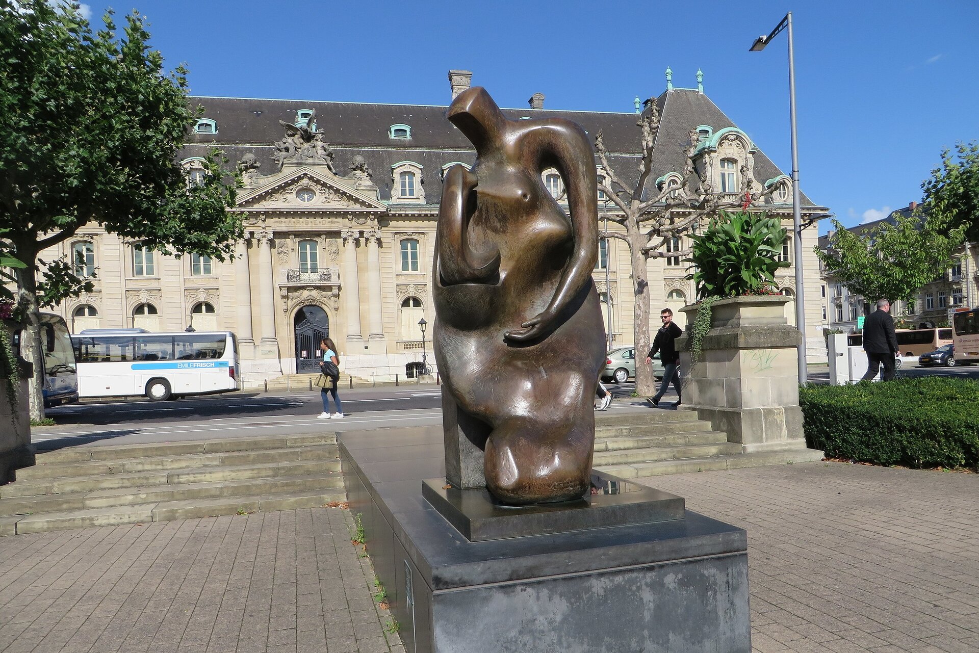 Zdjęcie przedstawia brązową rzeźbę. Rzeźba przedstawia matkę i dziecko. W tle znajduje się budynek oraz wyłożony kostką plac. Po obu stronach fotografii znajdują się krzewy oraz drzewa.