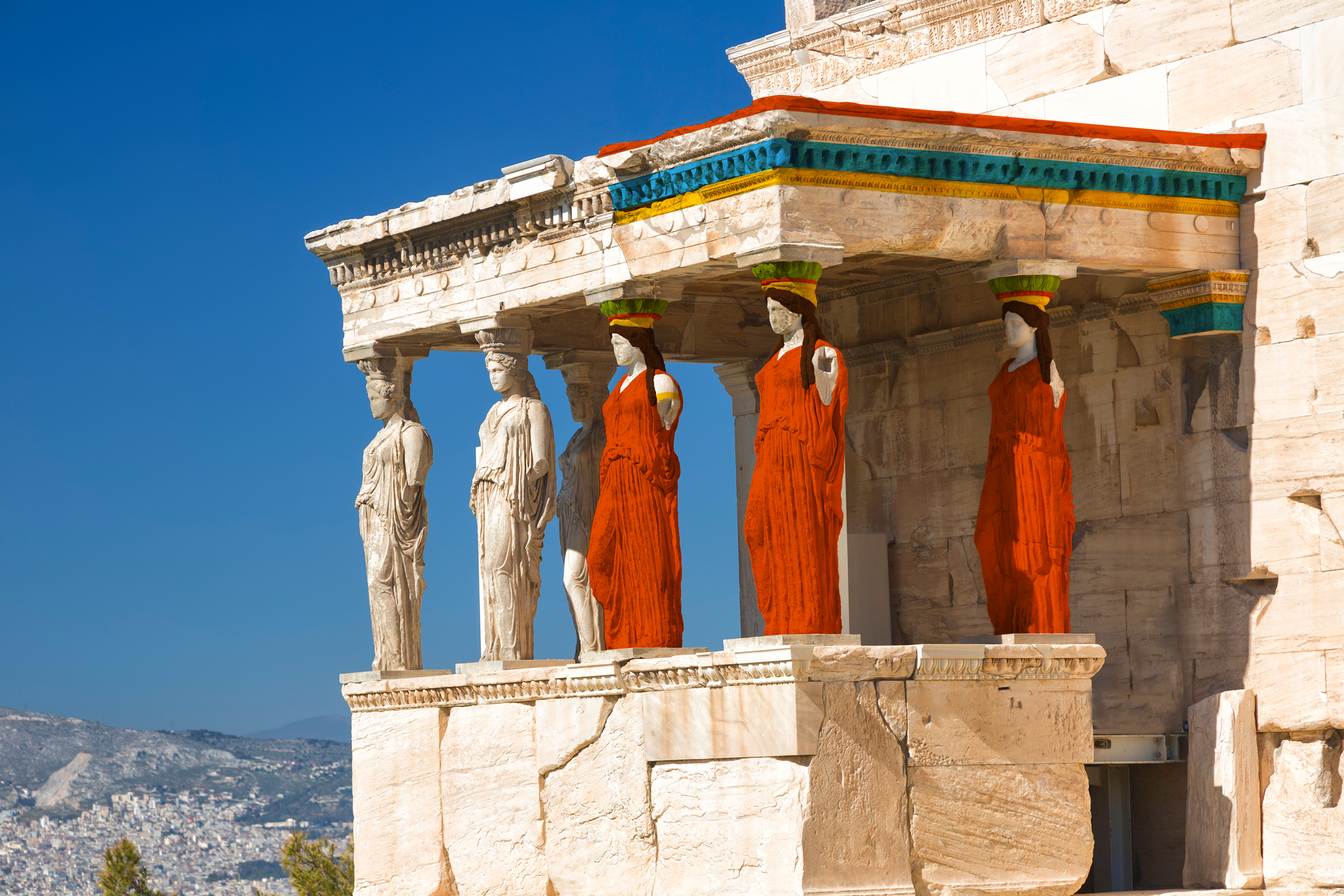 Fotografia przedstawia Erechtejon z widokiem od strony portyku. Fragment budowli jest po współczesnej rekonstrukcji. W centralnej jej części są kariatydy - kolumny w kształcie kobiet. Po lewej stronie fragment antyczny, zniszczony, w kolorze kamienia. Po prawej fragment zrekonstruowany, kolorowy. Kariatydy mają czerwone suknie, złote tarcze i ciemne włosy. Kapitele nad ich głowami oraz fryz są w kolorowe pasy. W tle ukazany jest krajobraz miasta i gór oraz bezchmurne niebo.