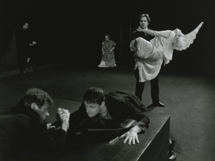 Na czarno‑białym zdjęciu przedstawiono fragment przedstawienia "Dziady - dwanaście improwizacji według Mickiewicza" reżysera Jerzego Grzegorzewskiego. Na pierwszym planie znajdują się mężczyźni, oparci o czarny stół, którzy siłują się na ręce. Ubrani są w czarne marynarki oraz spodnie. Za nimi stoi kolejny mężczyzna, również ubrany w czarną marynarkę i spodnie, który trzyma na rękach anioła w białej szacie. Na tle, w oddali, klęczy kobieta ubrana w ciemne szaty, a niedaleko niej stoi kolejny mężczyzna ubrany na czarno. Zdjęcie było zrobione na scenie.

