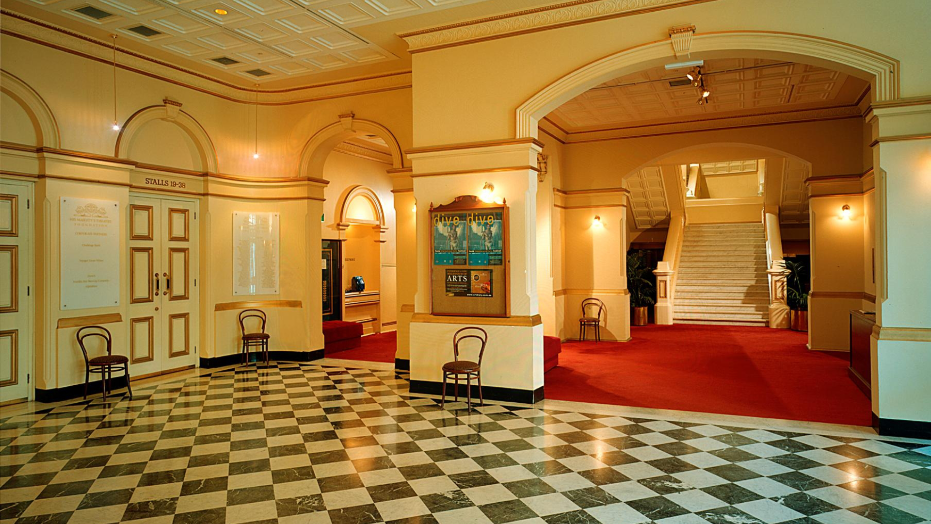 Zdjęcie przedstawia duże i przestronne foyer teatralne. Część pomieszczenia posiada dekoracyjną, błyszczącą terakotę. Dalsza część pokryta jest dywanem. Wnętrze jest wysokie i wzbogacone o dekoracyjne elementy architektoniczne. W foyer znajduje się kilka krzeseł, które stoją pod ścianami. Na ścianach znajdują się lampy - kinkiety, które oświetlają wnętrze.