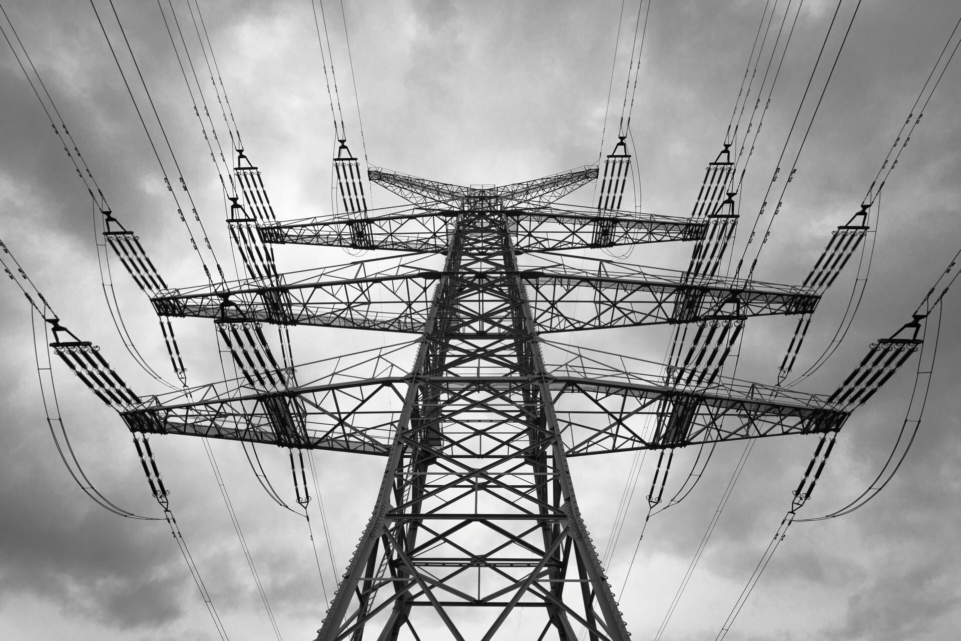 Zdjęcie czarno białe przedstawia słup wysokiego napięcia uwieczniony z żabiej perspektywy. Kilkudziesięciometrowej wysokości metalowa konstrukcja widoczna jest na tle pochmurnego nieba. Słup z obu stron dźwiga po sześć podwójnych przewodów energetycznych rozpiętych na specjalnych konstrukcjach z długimi układami izolatorów.
