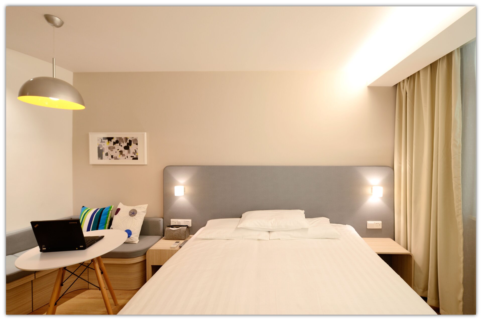 Ilustracja przedstawia sypialnię. Po prawej okno, obok którego znajduje się łóżko ze stolikami nocnymi po bokach. Po lewej kanapa narożna, obok której znajduje się stolik z laptopem. Nad stolikiem lampa sufitowa.