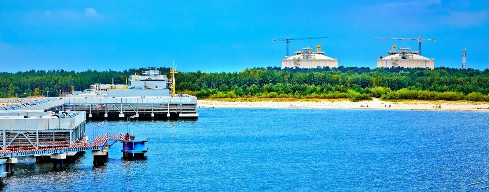 Zdjęcie gazoportu. Na pierwszym planie zbiornik wodny, po lewej infrastruktura gazoportu na morzu. Na drugim planie las i górujące nad nim dwa białe, duże zbiorniki na planie koła.