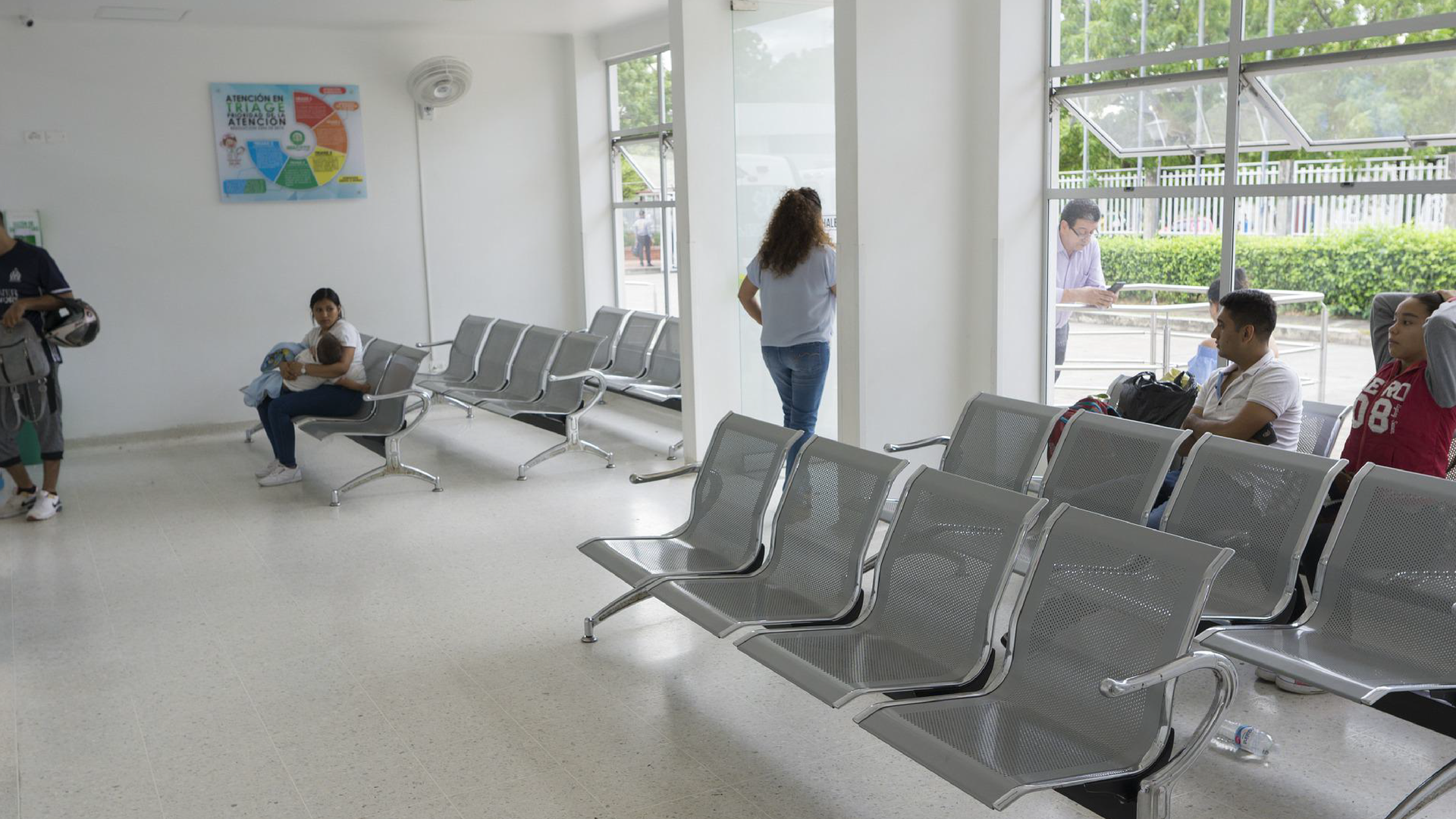 Zdjęcie przedstawia poczekalnię. W poczekalni ustawionych jest kilka rzędów siedzeń. Kilka osób siedzi na tych krzesłach. Widoczne jest również kilka osób, które stoją. 