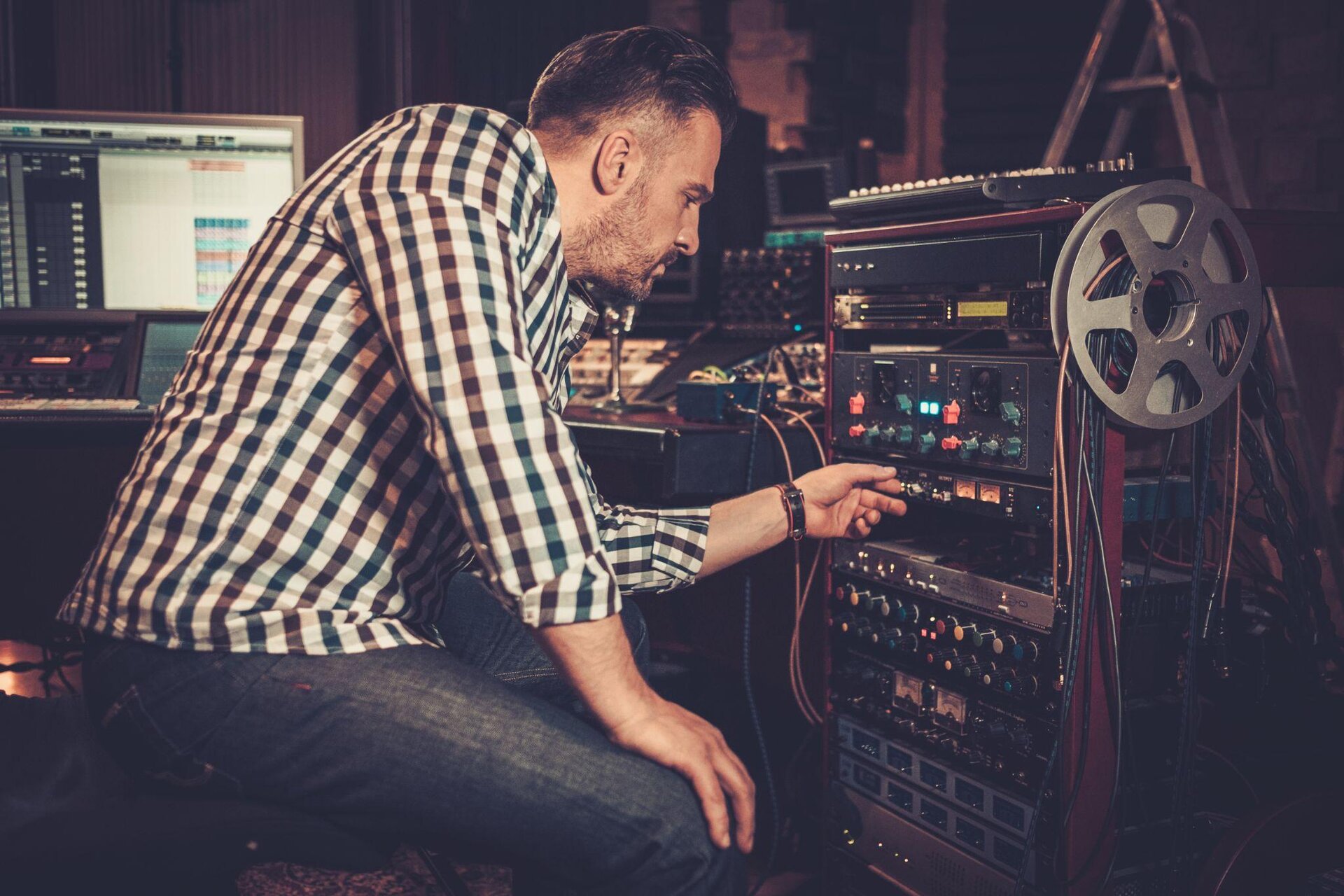 Na pierwszym planie fotografii znajduje się mężczyzna, który siedzi na czarnym krześle.  Ma na sobie białą koszulę w brązowo‑zieloną kratę oraz ciemnoniebieskie jeansy. Rękawy koszuli ma podwinięte na wysokości połowy przedramienia. Mężczyzna ma krótkie brązowe włosy, wąsy i brodę. Jego prawa dłoń dotyka prawego kolana. Na lewym nadgarstku ma czarny pasek. Po prawej stronie obrazka znajduje się kolumna z panelem sterowania. Jest na niej wiele kolorowych pokręteł. Jednego z nich dotyka mężczyzna swoją lewą dłonią. Z boku kolumny znajduje się szara szpulka, na której jest wiele kolorowych kabli. Na górze kolumny leży czarna konsola muzyczna z białymi przyciskami. Za mężczyzną znajduje się biurko z monitorem i konsolami. Z tyłu z prawej strony stoi drabina. Tło jest ciemnie, niewyraźne.