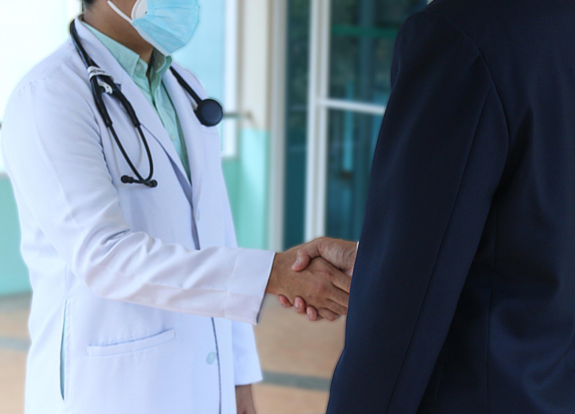 Zdjęcie przedstawia po lewej stronie lekarza ubranego w biały fartuch lekarski, z maseczką na twarzy oraz stetoskopem na szyi. Postać ma wyciągniętą prawą rękę i ściska dłoń osoby stojącej po prawej, ubranej w czarny garnitur.