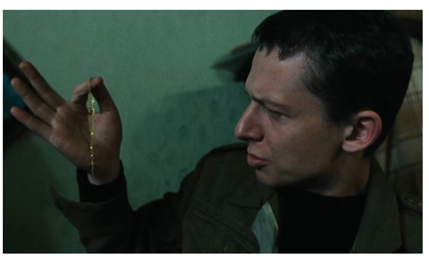 Fotografia przedstawia mężczyznę w sile wieku. Postać ukazuje lewy profil. Mężczyzna ma wysokie czoło, zmarszczone brwi, zmrużone oczy i duży nos. Lekko wysuwa dolną wargę. Jego ciemne włosy są krótko ostrzyżone i proste. Prawą rękę unosi do wysokości oczu. Na palcu wskazującym ma przewieszony cienki, złoty łańcuszek. Mężczyzna jest ubrany w kurtkę z rozpiętymi mankietami i koszulkę. W tle ukazana jest ściana z wzorzystą tapetą.