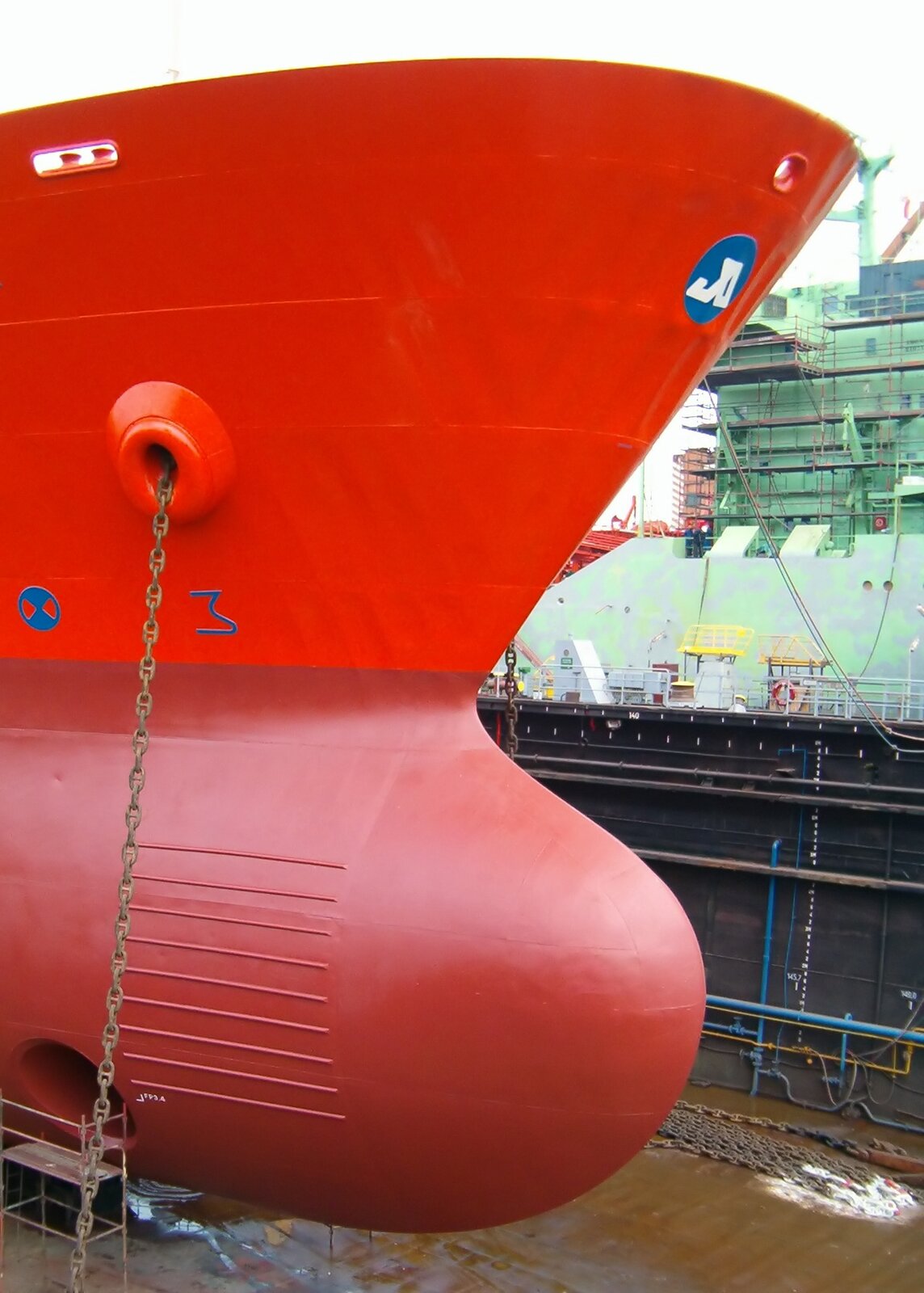 Fotografia przedstawia dziób okrętu, który został pomalowany na kolor czerwony w dwóch odcieniach: góra w kolorze jasnoczerwonym, a dół w ciemnoczerwonym. Kolorem niebieskim naniesiono symbole oraz logo armatora. Dziób okrętu przebywa w suchym doku. W tle widoczny jest inny budowany okręt.