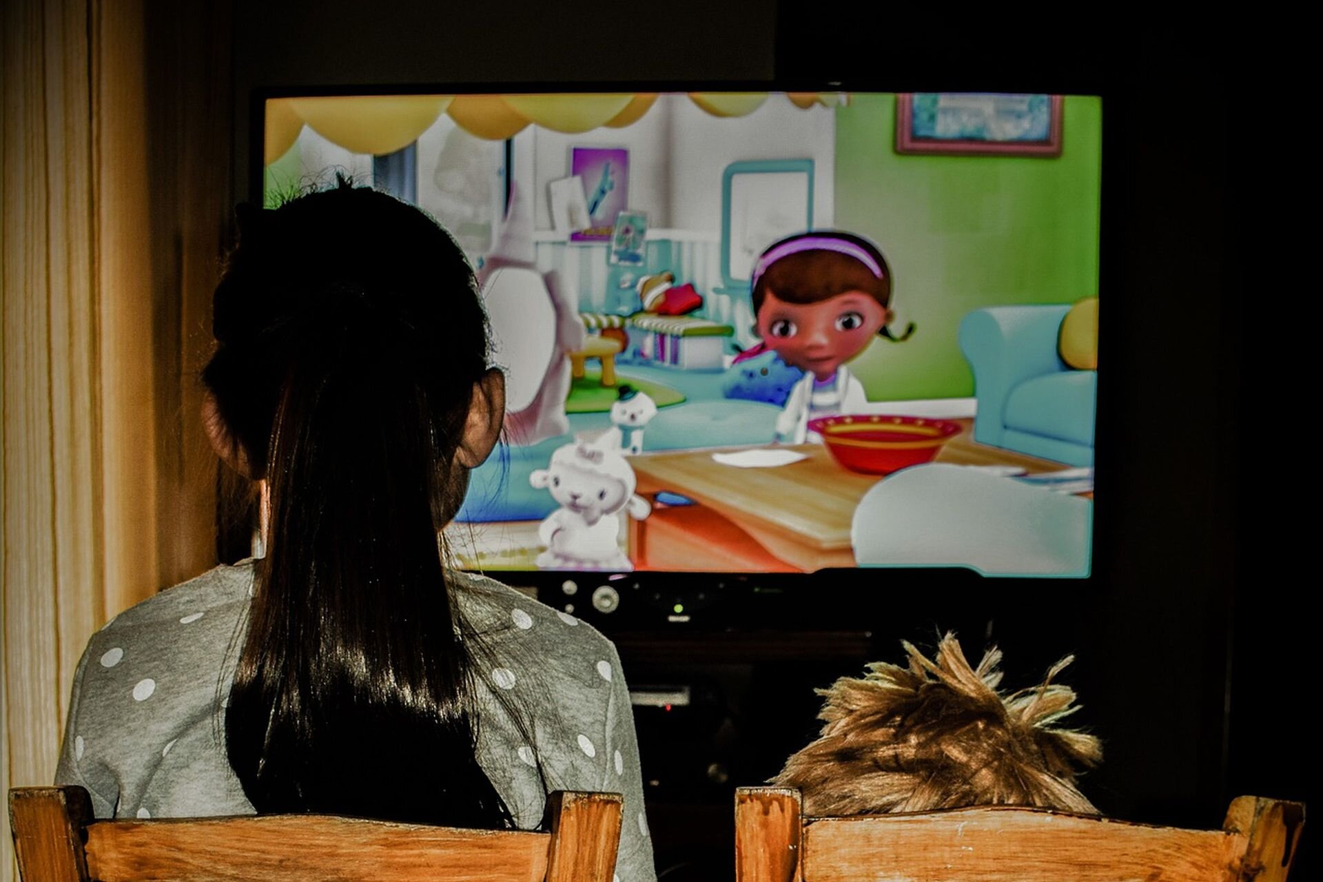 Ilustracja przedstawia dziewczynkę, która ogląda bajkę, siedząc przed ekranem telewizora.