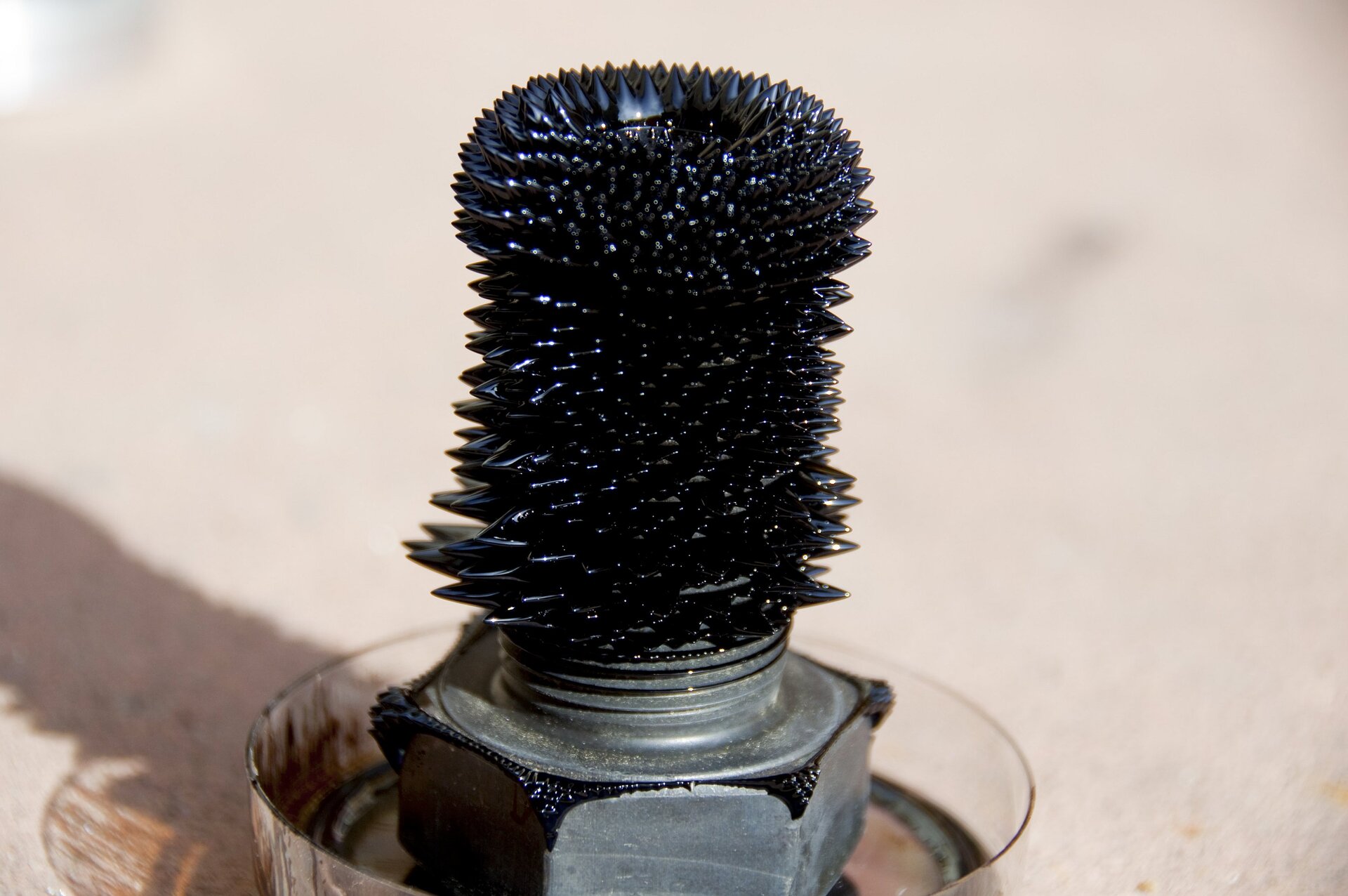 Fotografia przedstawia walcowaty ciemny magnes oblepiony opiłkami metalowymi. Tło jasne.