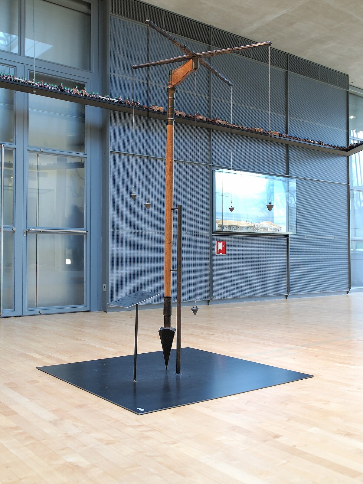 Fotografia przedstawia wnętrze muzeum, w którym zaprezentowano rekonstrukcję rzymskiego narzędzia mierniczego. Narzędzie jest dosyć wysokie, zbudowane z metalowego grotu, drewnianego trzonka, u góry którego zamontowano dwie metalowe belki dociążone metalowymi odważnikami. 