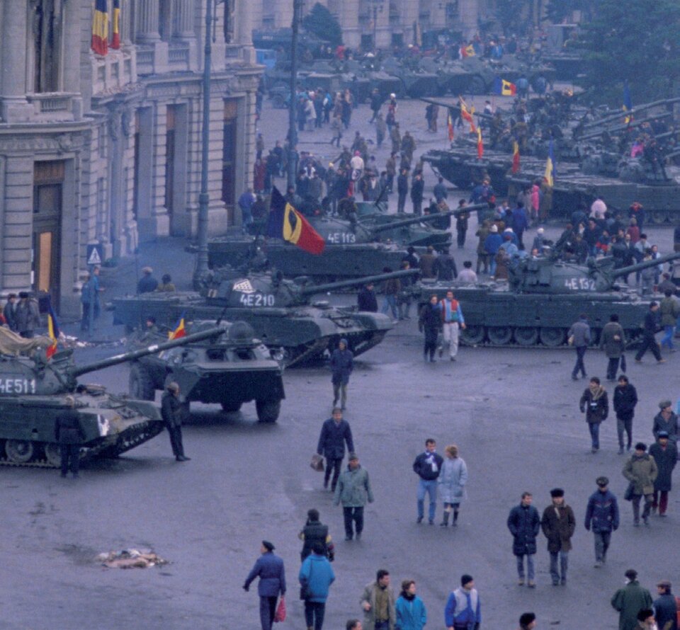 Zdjęcie przedstawia miejski plac, na którym stoi wiele czołgów z rumuńskimi flagami. Pomiędzy czołgami spacerują ludzie. Po lewej stronie stoi kamienica. Nad czołgami unosi się słabo widoczny dym.