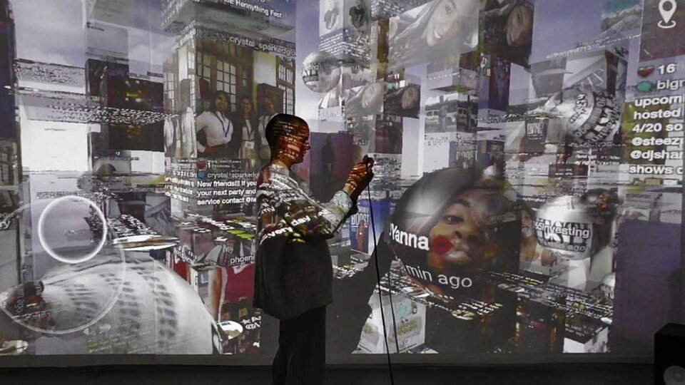 Prostokątna fotografia ukazuje kobietę o krótkich włosach. Ubrana jest w szary sweter oraz czarne spodnie. Znajduje się w pomieszczeniu, w którym wyświetlone zostały prezentacje multimedialne. Są one wykonane w stonowanych barwach.