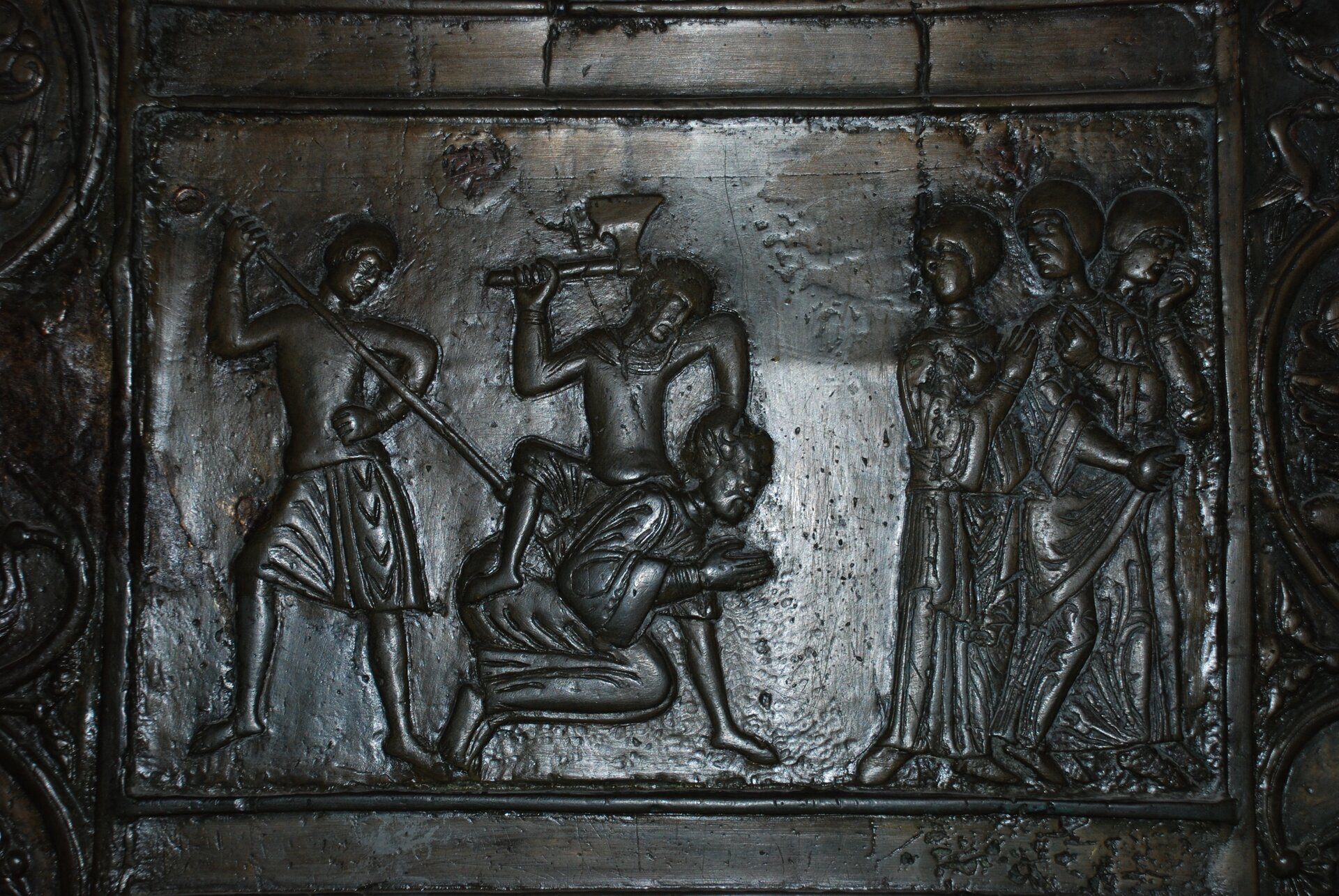 Fotografia przedstawia Drzwi Gnieźnieńskie, scena męczeńskiej śmierci św. Wojciecha. Osoba jest poddawana torturom. Tablica na foto wygląda na wykonaną z metalu i ciężką.