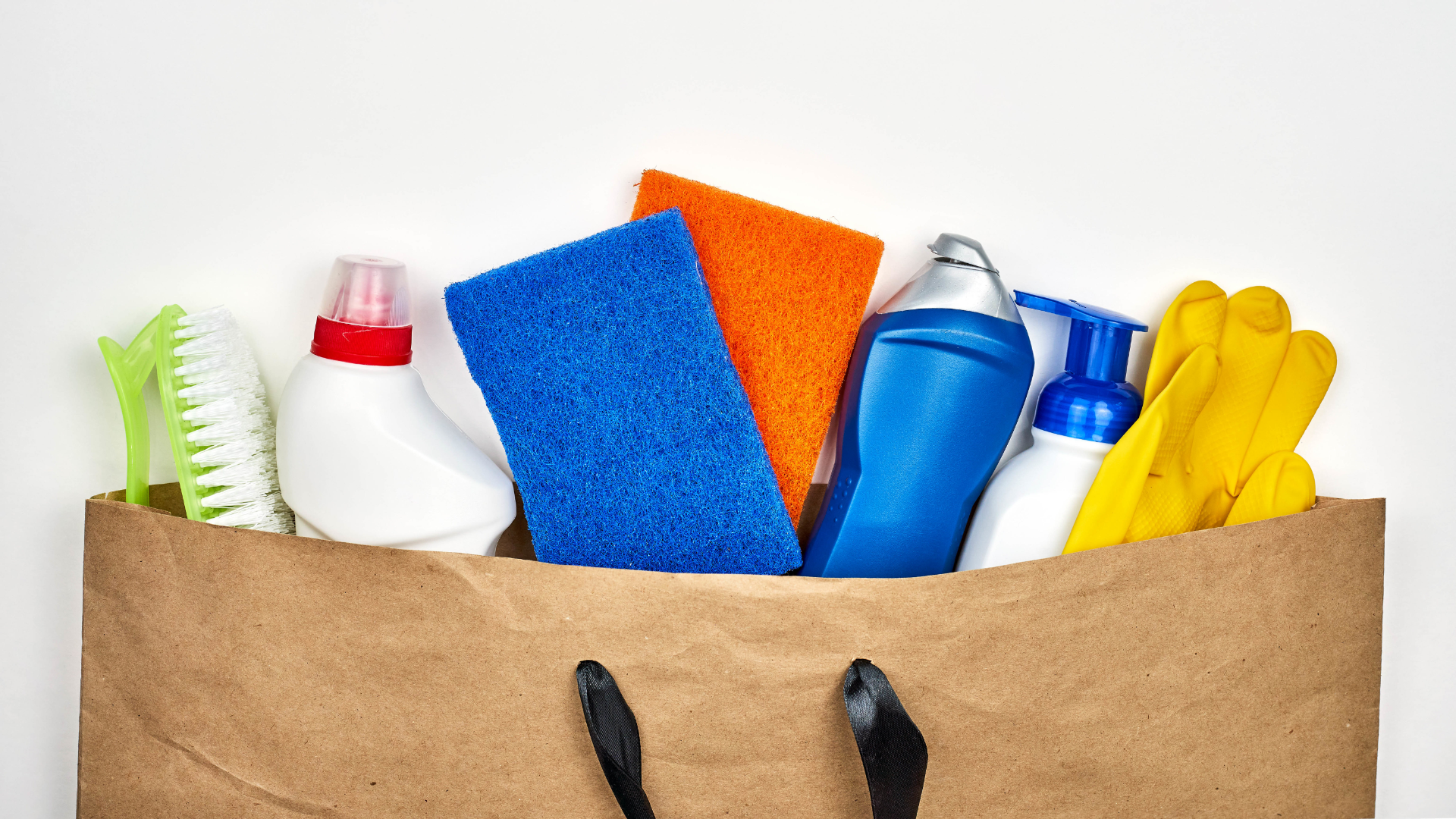 Zdjęcie przedstawia papierową torbę ze środkami czyszczącymi. Widoczne są: gumowe rękawice, butelki ze środkami czyszczącymi, gąbki oraz szczotka.