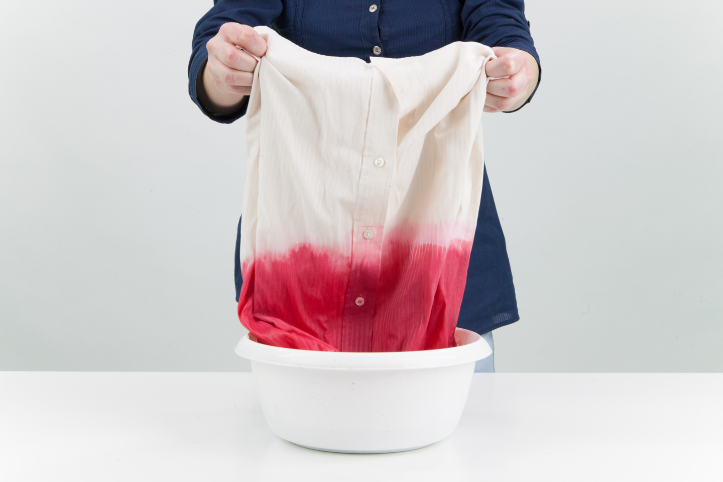 Fotografia przedstawia osobę farbującą ubranie w  białej miednicy. Jest to biała koszula zafarbowana do połowy na czerwono.
