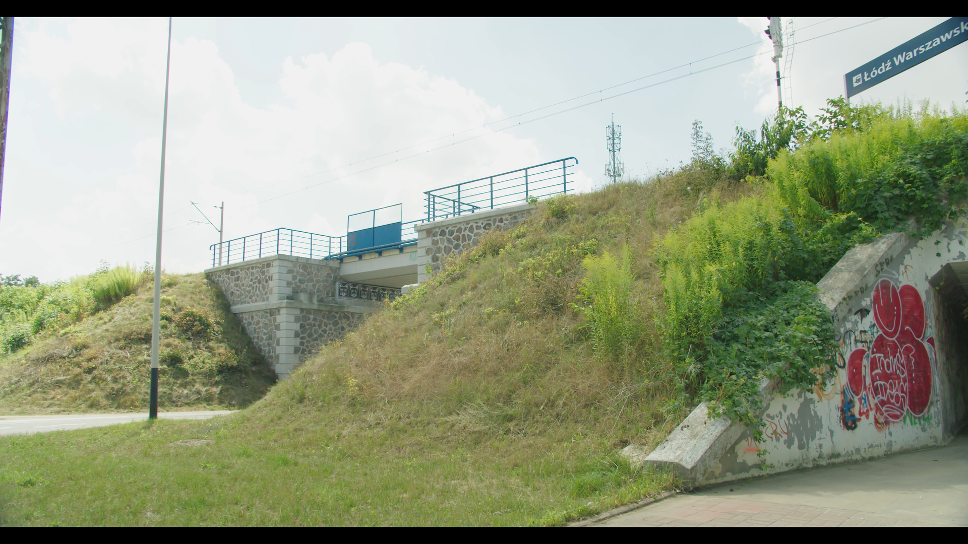 Ilustracja przedstawia przejście pod torami. Widoczny jest nasyp kolejowy z wiaduktem ponad drogą. Po prawej stronie zdjęcia widać wejście do przejścia pod nasypem.