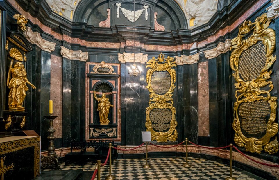 Ilustracja przedstawia zdjęcie wnętrza Kaplicy Wazów na Wawelu w Krakowie. Wnętrze jest bogato zdobione różnorodnymi kolorowymi (czarny, różowy, biały) materiałami: wapieniami, alabastrem oraz złotem. W niewielkich niszach znajdują się złote posągi, wszystkie ściany pokryte są okładzinami oraz złoconymi sztukateriami. Na środku ściany ołtarzowej znajduje się oprawione w złote ramy malowidło. Między ścianami a tamburem biegnie wydatny gzyms okalający całe wnętrze.