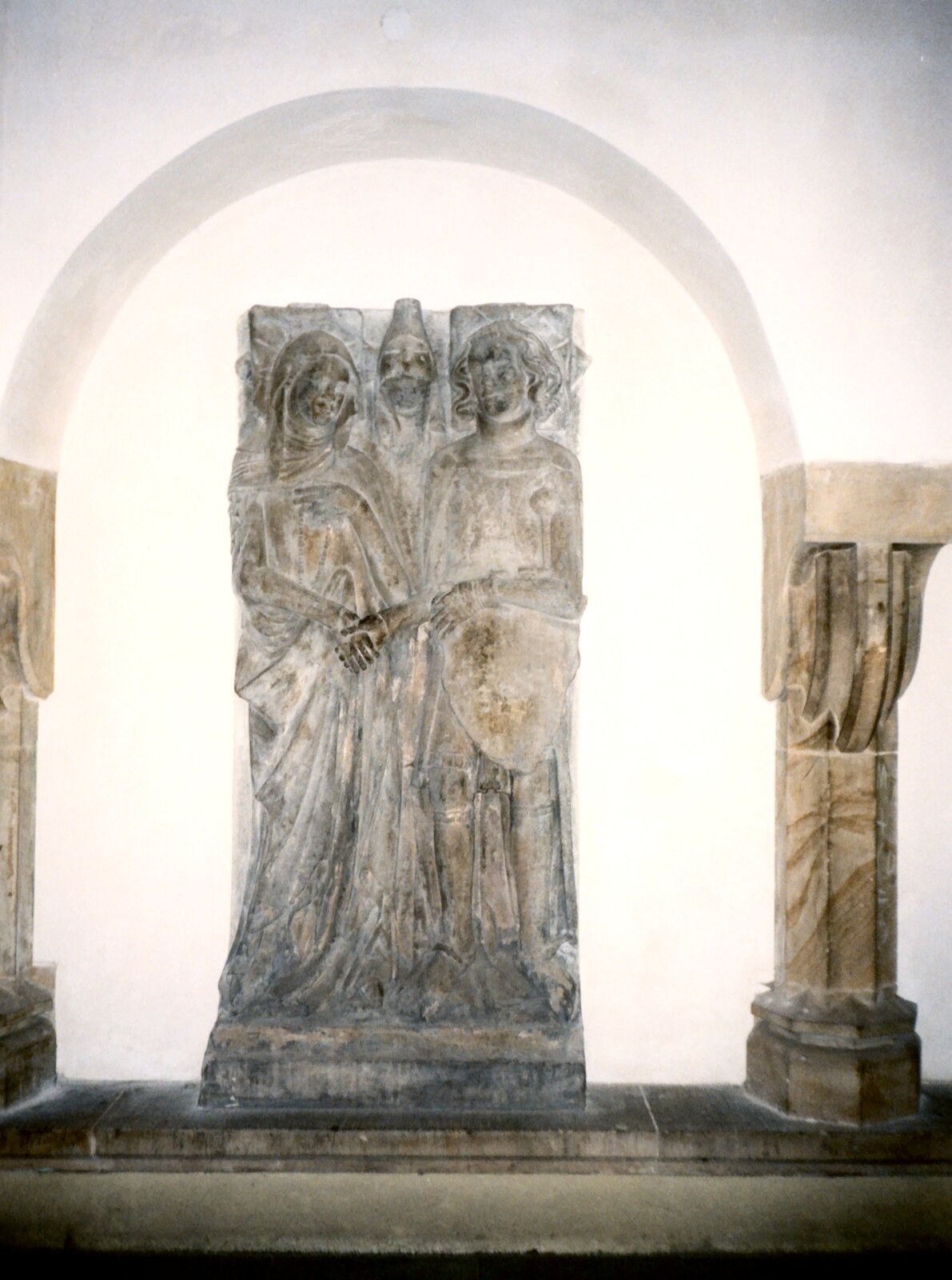 Fotografia przedstawia płytę nagrobną, na której przedstawione zostały dwie postacie - kobiety i mężczyzny - w strojach charakterystycznych dla średniowiecza. Płyta została wyeksponowana w pozycji pionowej na tle białej ściany.