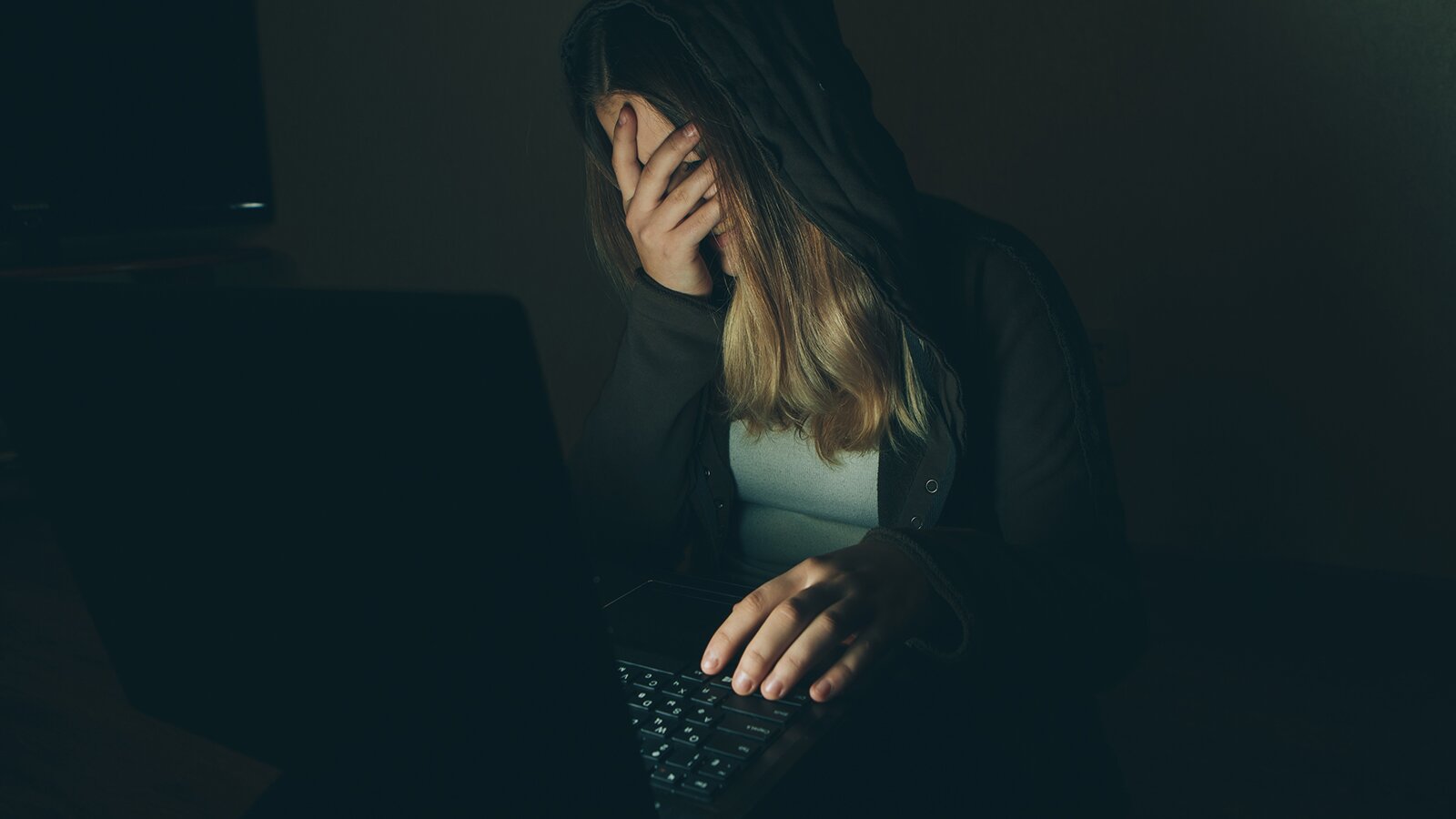 Zdjęcie przedstawia siedzącą młodą dziewczynę, która ubrana jest w czarną bluzę z kapturem i zielonkawą bluzkę. Ma długie, rozpuszczone blond włosy, na które ma założony kaptur bluzy. Dziewczyna siedzi przed monitorem laptopa, ma lekko opuszczoną głowę. Lewą dłoń trzyma na klawiaturze, a prawą dłonią zakrywa twarz.