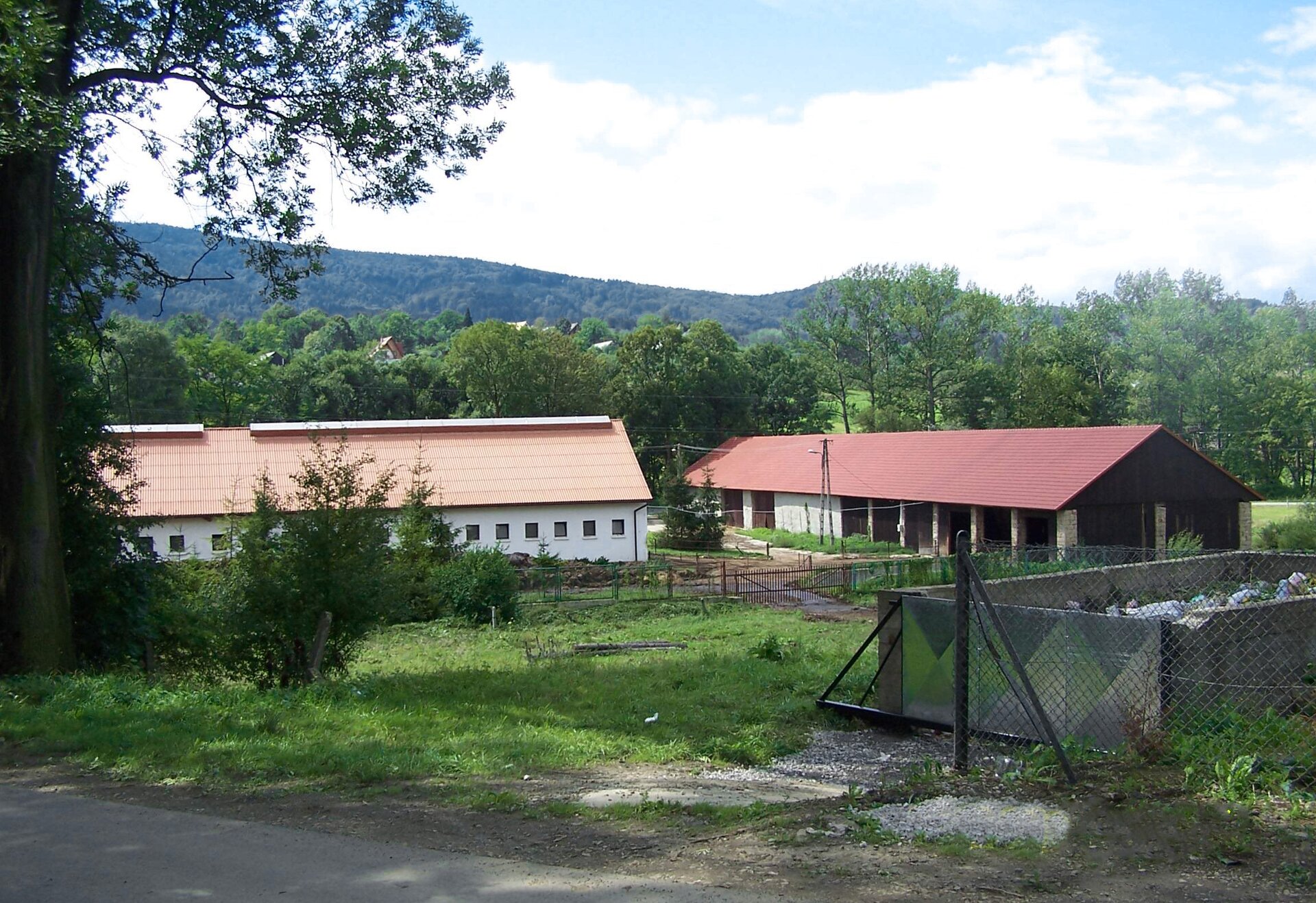 Zdjęcie przedstawia Państwowe Gospodarstwo Rolne. Na pierwszym planie znajduje się trawnik oraz kontener na odpady. Dalej jest ogrodzenie. Za nim znajdują się zabudowania. Budynki są niskie oraz długie. Posiadają wiele okien.