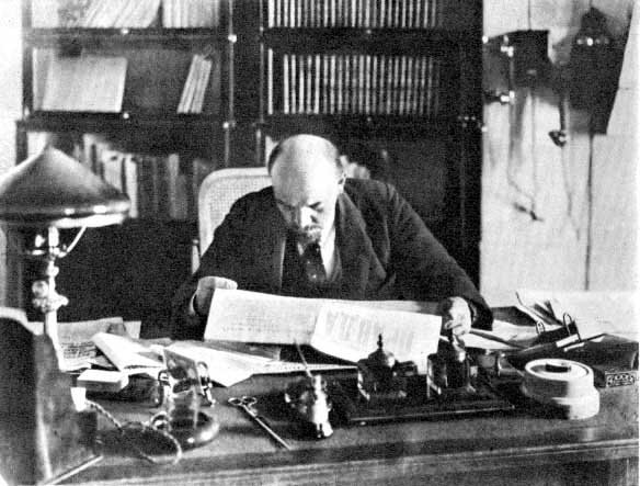 Kliknij, aby powiększyć Zdjęcie przedstawia pomieszczenie, w którym za biurkiem siedzi Włodzimierz Lenin i czyta gazetę. Na biurku są kartki, lampka, przybory do pisania. Za biurkiem widać regał z książkami.
