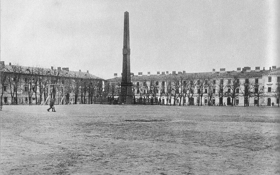 Ilustracja przedstawia duży plac, na którym stoi pomnik. Pomnik jest w kształcie zwężającego sie ku górze wąskiego prostopadłościanu. Wokół pomnika rosną drzewa. Plac otoczony jest w dwóch stron zabudowaniami. Zabudowania to ciąg budynków trzykondygnacyjnych, pokrytych dwuspadowym dachem.