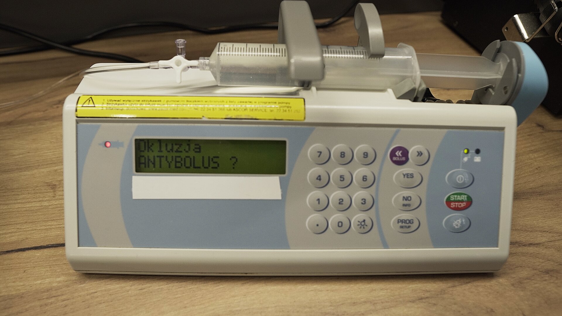 Na zdjęciu strzykawka wypełniona wodą znajduje się w ramieniu pompy infuzyjnej. Na ekranie aparatu wyświetlana jest informacja: "Okluzja, Antybolus?".