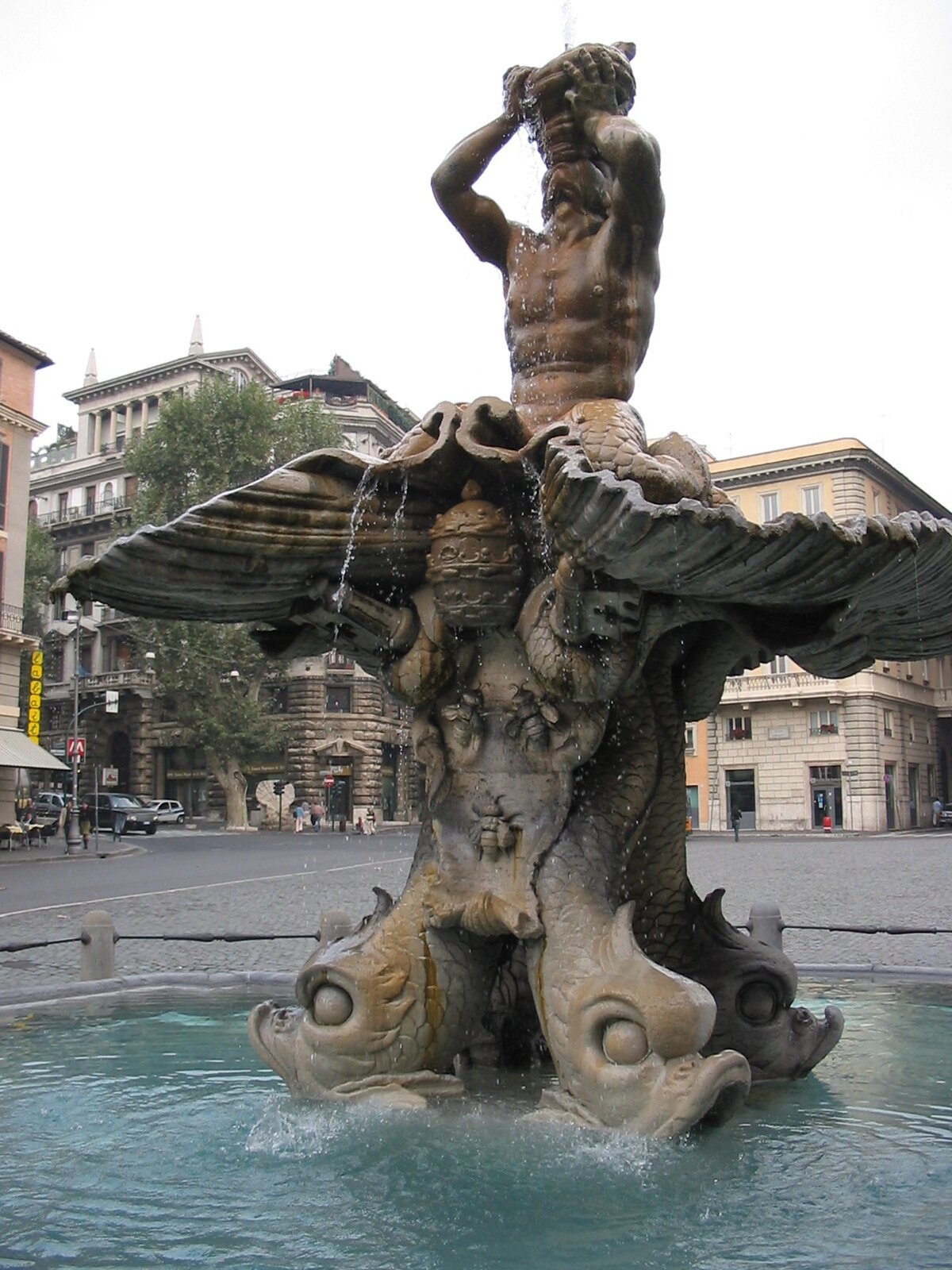 Ilustracja przedstawia zdjęcie czynnej fontanny Trytona w Rzymie. Rzeźba przedstawia bóstwo morskie, które trzyma róg, z którego wypływa woda. W podstawie fontanny znajdują się podobizny delfinów, które podtrzymują muszlę, na której umieszczona jest postać Trytona. Fontanna została wkomponowana w plac, wokół którego widać budynki.