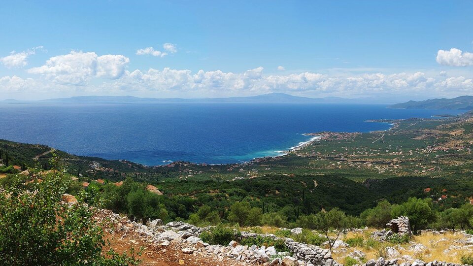 Kolorowa fotografia przedstawia panoramiczne ujęcie z góry nadmorskiego krajobrazu. Na tle niebieskiego nieba widać białe chmury, poniżej rozciąga się morze okolone wysokimi górami. Na wprost znajduje się zielona, nadmorska dolina z łąkami i domami ludzi. Domki są malutkie, nie widać ich dokładnych kształtów.