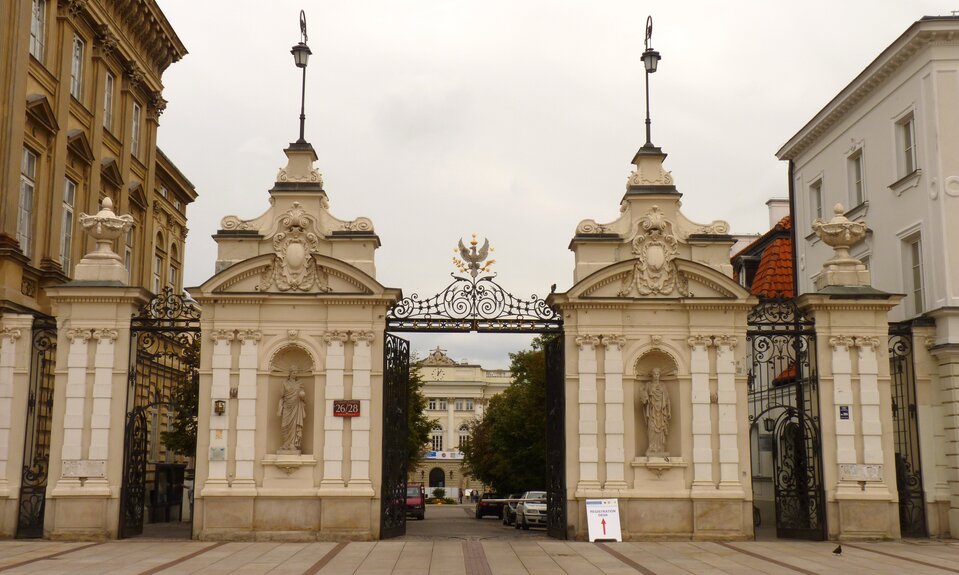 Fotografia przedstawia okazałą bramę. W jej krańcach stoją ozdobne kolumny. Na ich szczytach stoją rzeźby w kształcie waz ozdobionych girlandami z liści. Bliżej środka znajdują się metalowe furtki. Po obu stronach otwartej brany stoją dwie szerokie kolumny z wnękami. We wnękach umieszczone są rzeźby wyobrażające kobiety ubrane w obszerne togi. Postać stojąca po lewej trzyma w prawej dłoni zwój pergaminu, a w lewej dość dużą kulę. Kobieta stojąca po prawej stronie trzyma w prawej dłoni hełm. Pośrodku bramy umieszczony jest orzeł z rozpostartymi skrzydłami. Zaciska szpony na złotych gałązkach. Na głowie ma złotą koronę. Wokół ptaka umieszczono pięć pięcioramiennych gwiazd: nad koroną, obok skrzydeł, obok szponów. Pod orłem znajduje się napis: Uniwersytet Warszawski.