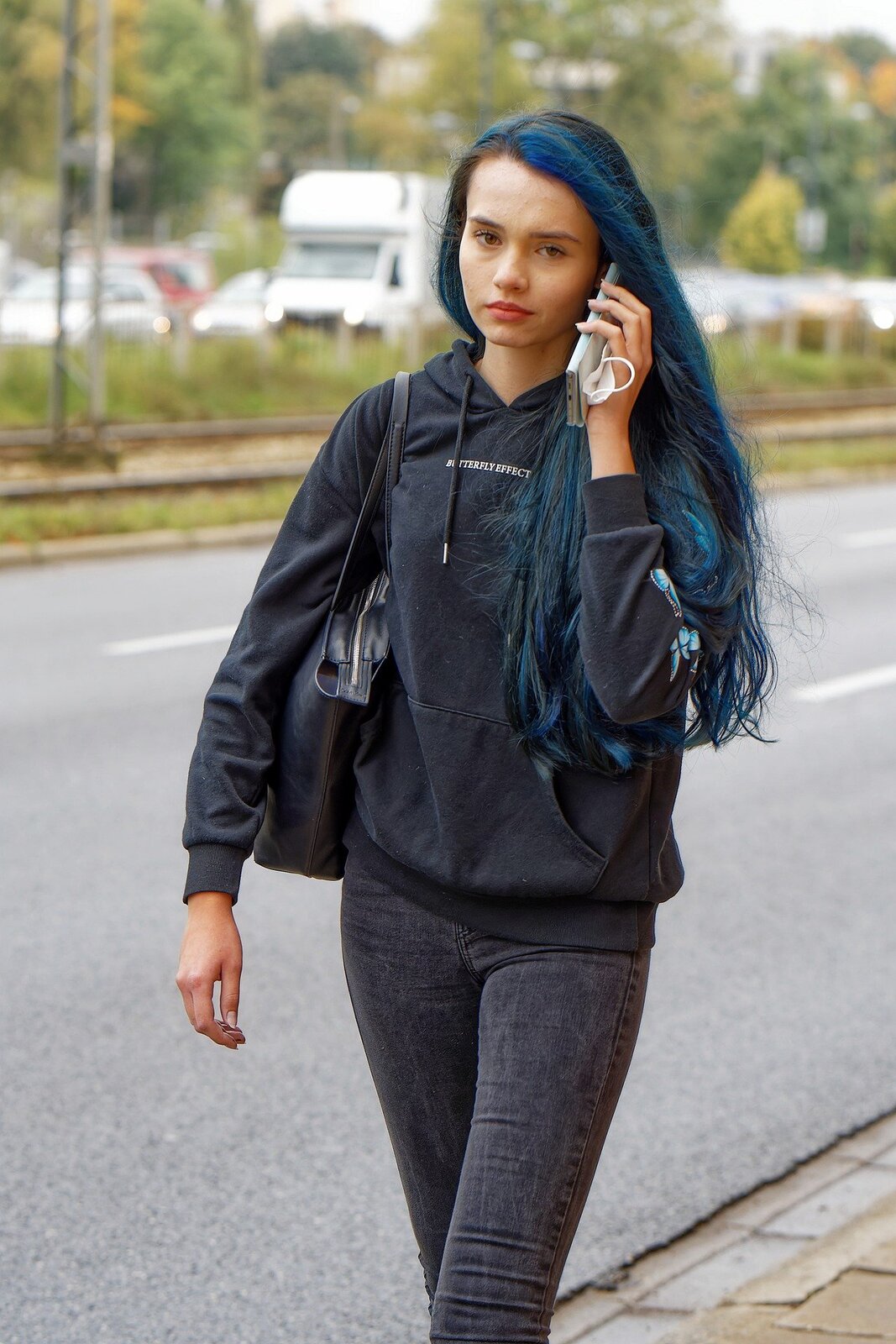Kolorowa fotografia przedstawia młodą dziewczynę, która idzie ulicą i rozmawia przez telefon komórkowy. Dziewczyna ma delikatny makijaż, ma wydatne naturalnie czerwone usta i proste, czarne, długie włosy z pasmami pofarbowanymi na niebiesko. Ubrana jest w czarną bluzę z białym napisem z przodu, niebieskimi motylami na lewym rękawie i grafitowe jeansy. Na prawym ramieniu ma zawieszoną czarną torebkę, w lewej ręce oprócz telefonu trzyma też maseczkę do zasłaniania twarzy. W tle widać ulicę, tory tramwajowe, jadące samochody i rosnące nieopodal drzewa. 