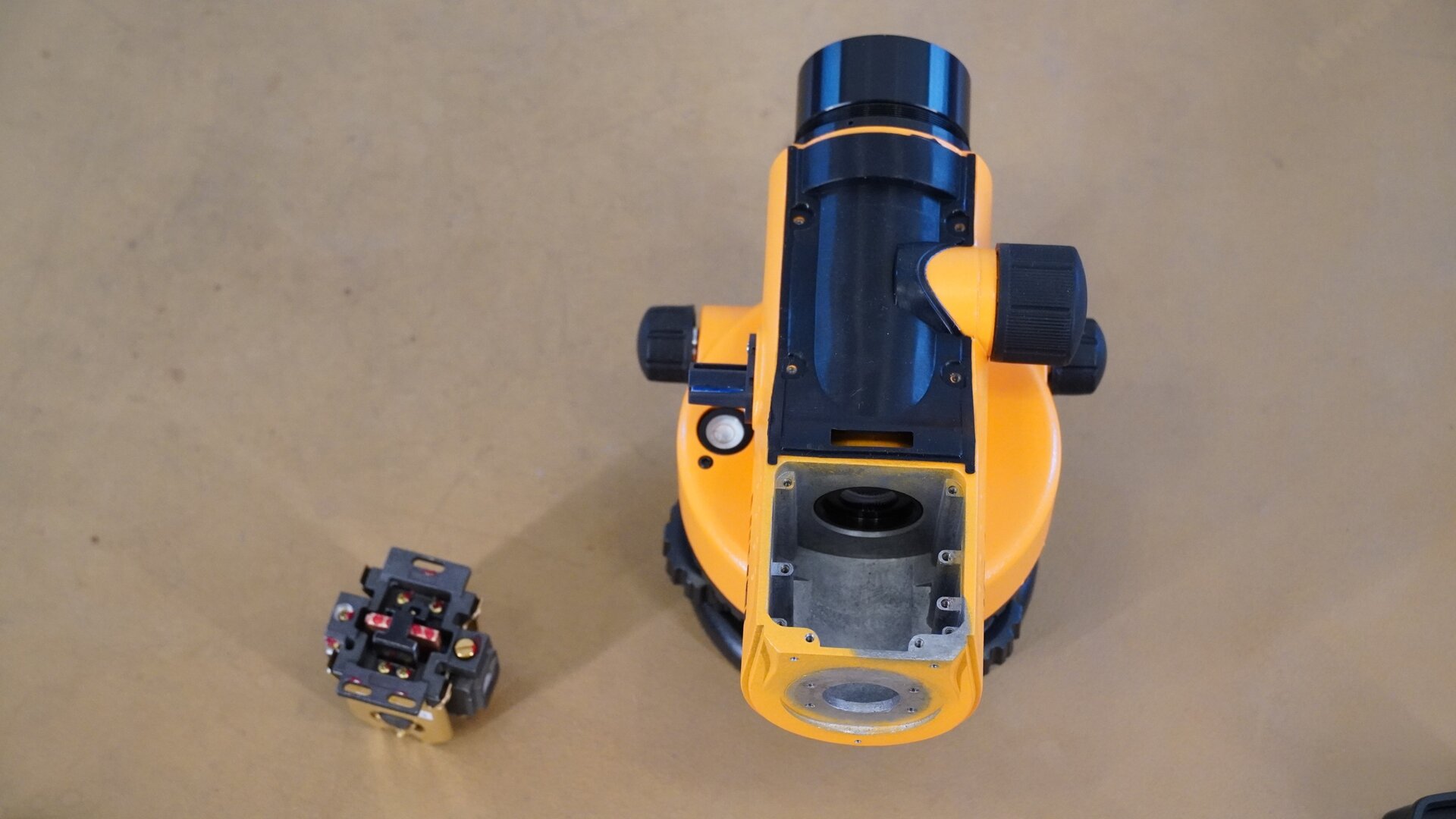 Zdjęcie przedstawia widok niwelatora z góry, po zdjęciu górnej osłony. Tuż za okularem widoczne jest puste miejsce, w którym był umieszczony kompensator. Kompensator znajduje się teraz obok niwelatora. Dalej widoczny jest tubus zakończony obiektywem, a po bokach pokrętła.