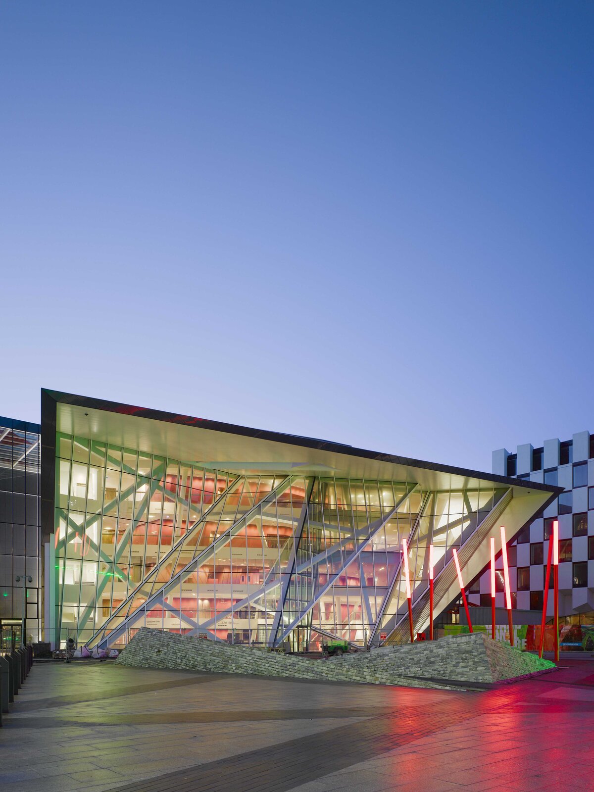 Ilustracja przedstawia budynek Bord Gáis Energy Theatre w Dublinie. Teatr ma geometryczny, nieregularny kształt. Cała przednia ściana jest przeszklona, składa się z podłużnych kawałków szkła. Nie jest ona płaska, ale tworzy wypukłe wzory i przypomina harmonijkę. Obramowanie teatru z przodu tworzą czarne kafelki. Zaraz przed budynkiem, na zewnątrz znajdują się dwie murowane instalacje o kształcie ramp. Na jednej z nich są poustawiane cienkie czerwone słupki z czerwonym światłem na szczycie. Wewnątrz, przymocowane do szyb znajdują się białe kolumny, poprzecinane jedna drugą. Dalej znajdują się białe kondygnacje teatru, schody, drzwi do sal. Ze środka budynku dochodzi światło. Fotografia została zrobiona o zmierzchu lub brzasku, w słabym świetle.