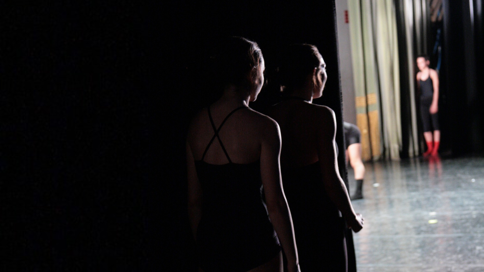 Zdjęcie zrobione jest zza kulis teatralnych. Na pierwszym planie znajdują się sylwetki dwóch kobiet, które obserwują, to co dzieje się na scenie. Zarówno panie, jak i całe kulisy pozostają w mroku. W tle widoczna jest scena i dwie osoby na scenie.