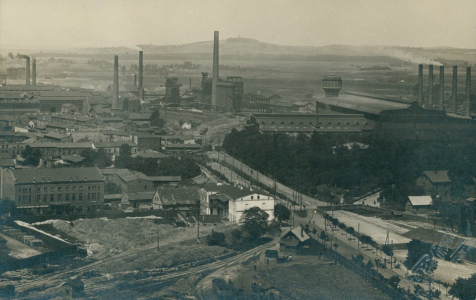 Zdjęcie przedstawia panoramę górniczego miasta. Ukazane są liczne zabudowania fabryczne, hale, kominy. Prowadzi do nich szeroka droga. W tle niewielkie wzgórza.
