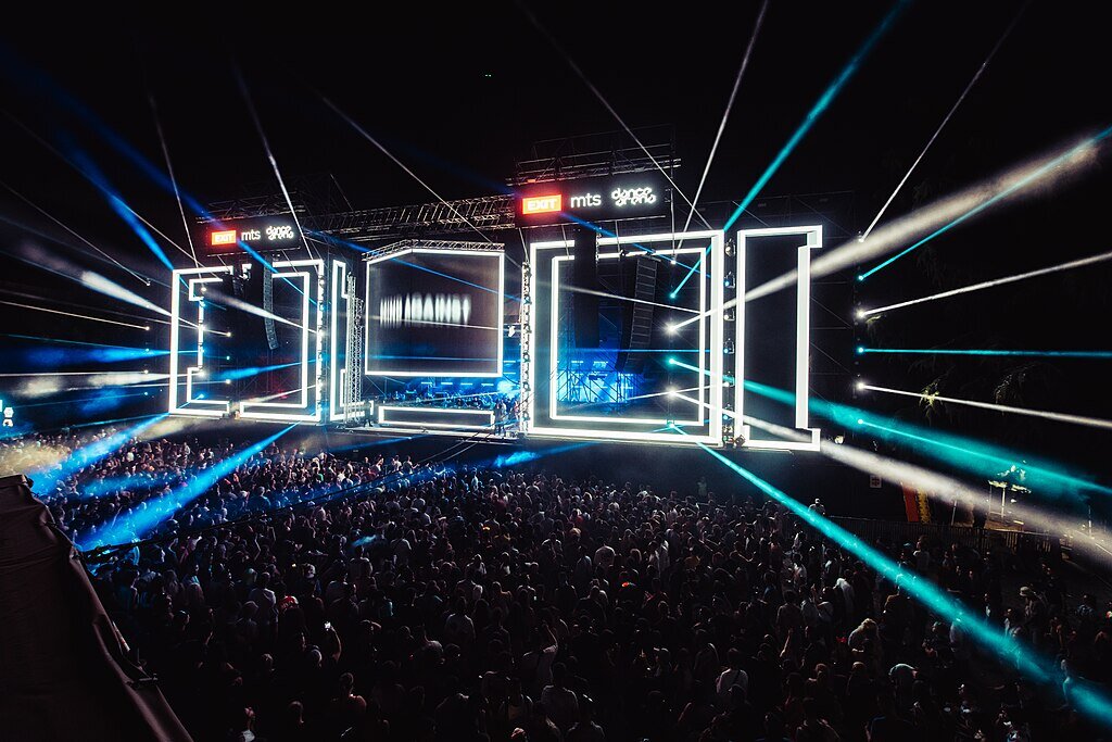 Zdjęcie przedstawia scenę koncertową oraz tłum widzów. Scena składa się z wielkich ram, zbudowanych ze świecących elementów, z których rozchodzą się strumienie ostrego światła w stronę widowni. Dookoła zalega ciemność.