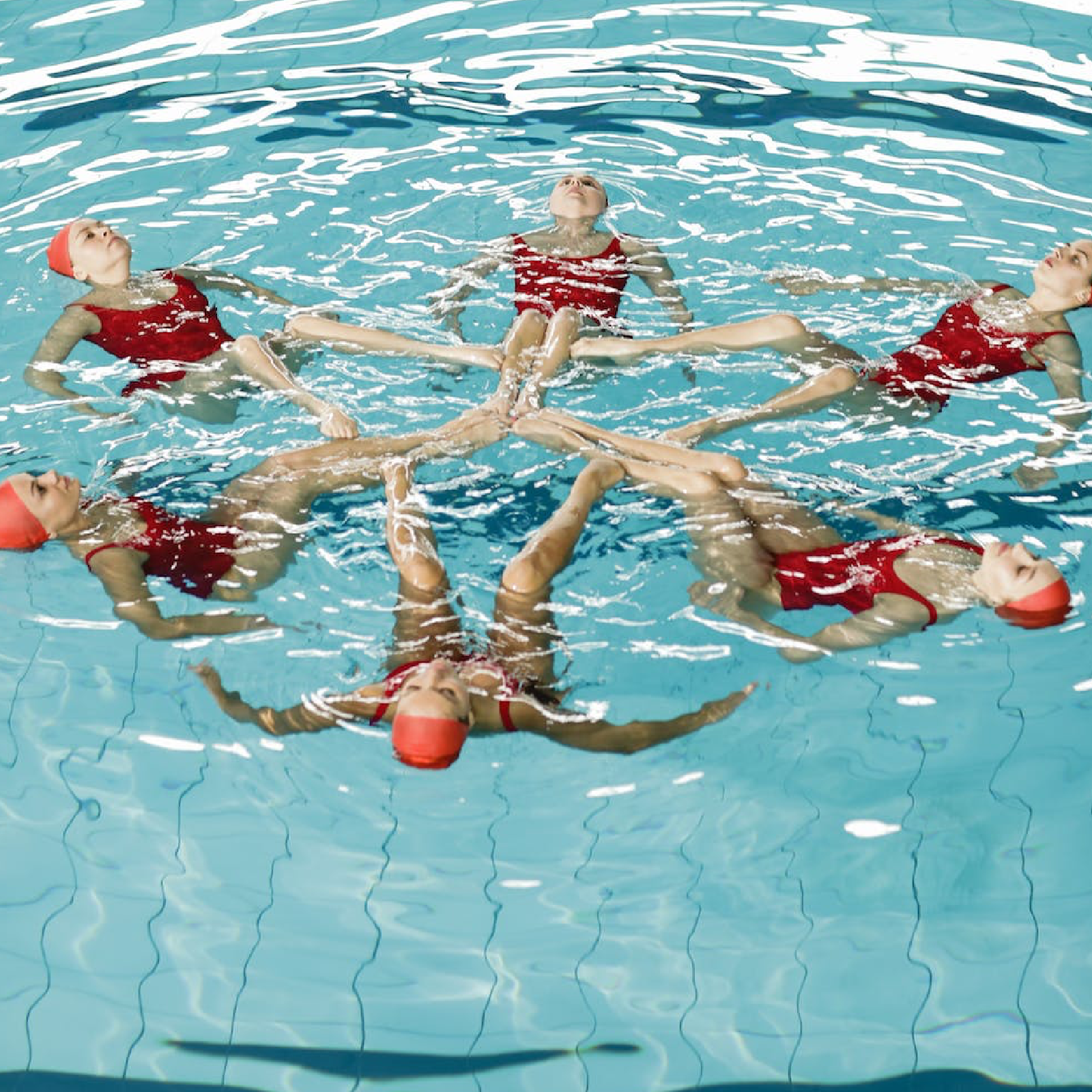 Ilustracja przedstawia sześć kobiet w czerwonych strojach kąpielowych i czepkach. Unoszą się na powierzchni wody, leżąc na plecach. Tworzą ze swoich ciał gwiazdę, stykając się stopami.