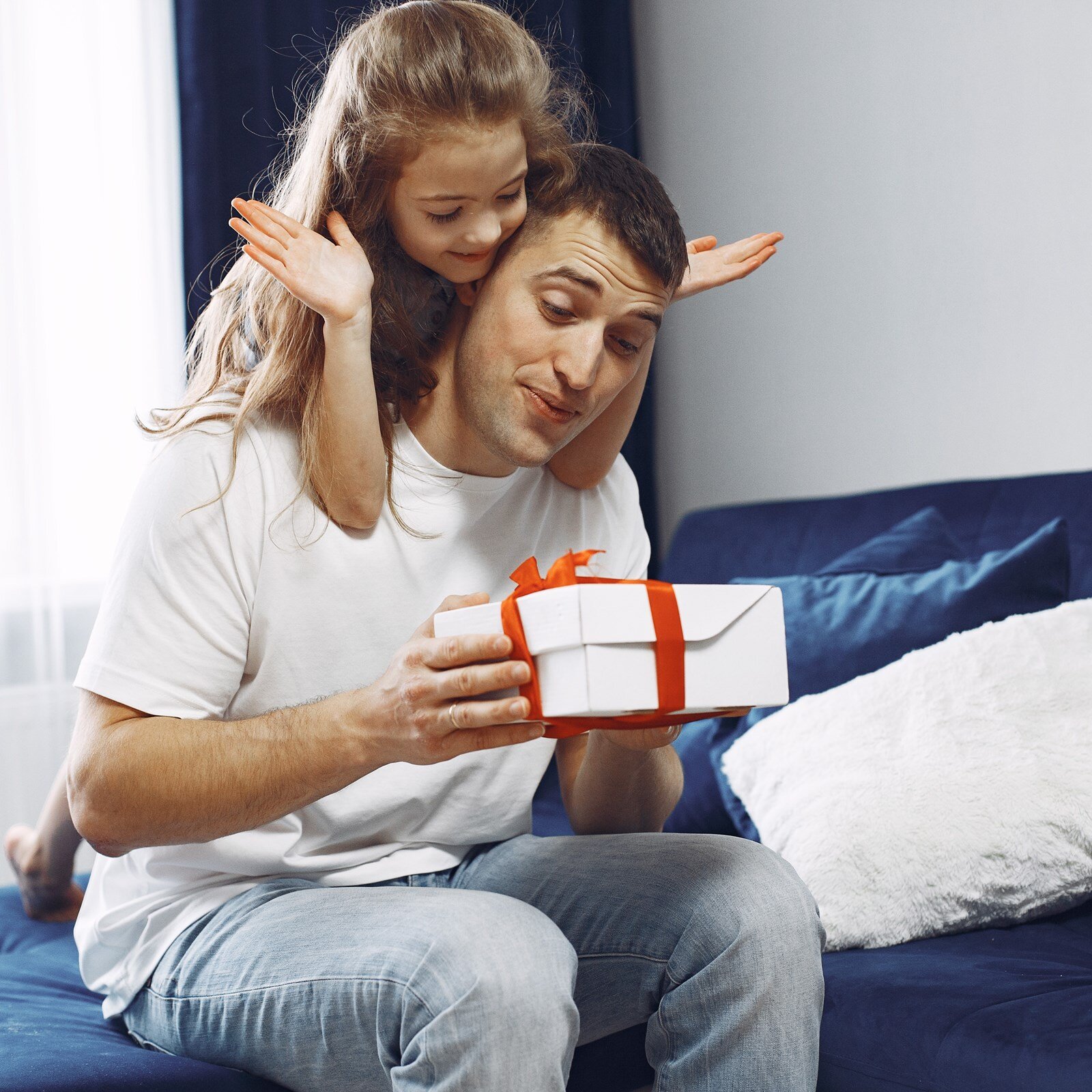 Zdjęcie przedstawia mężczyznę siedzącego na brzegu łóżka i trzymającego w dłoni pudełko obwiązane czerwoną wstążką. Nad głową widoczna mała dziewczynka opierająca się o jego plecy. 