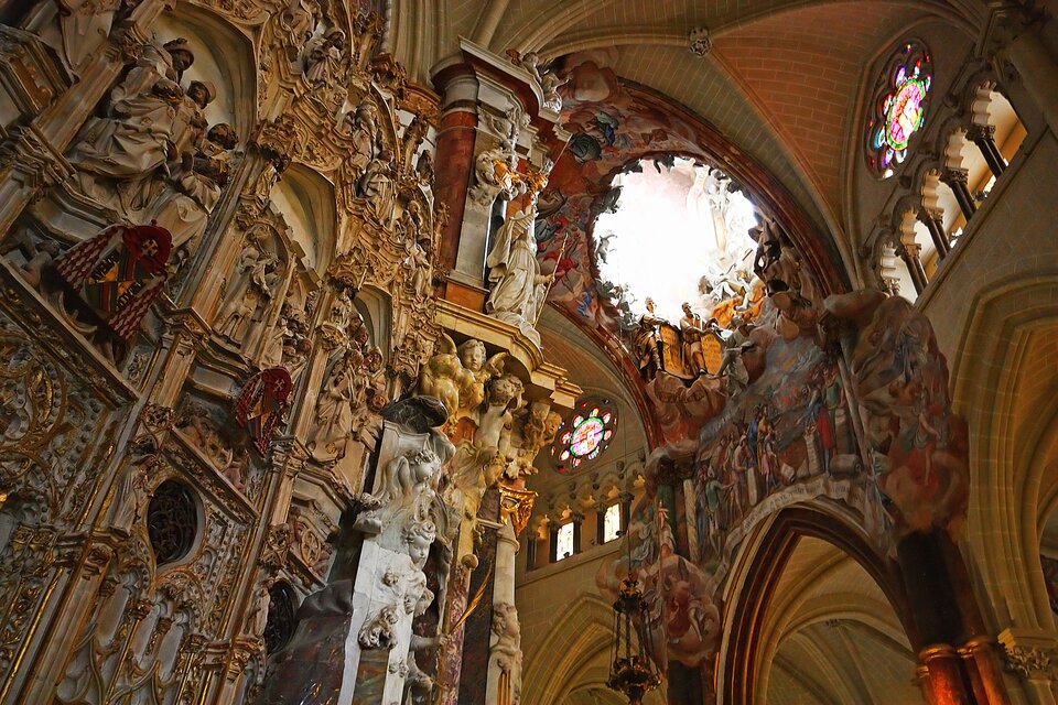 Zdjęcie przedstawia wnętrze katedry w Toledo. Na ścianie sklepienia znajdują się kolorowe freski. Przy filarze i we wnęce znajdują się rzeźby figur. Głowice kolumn oraz nastawa ołtarza są ozdobione złotem.