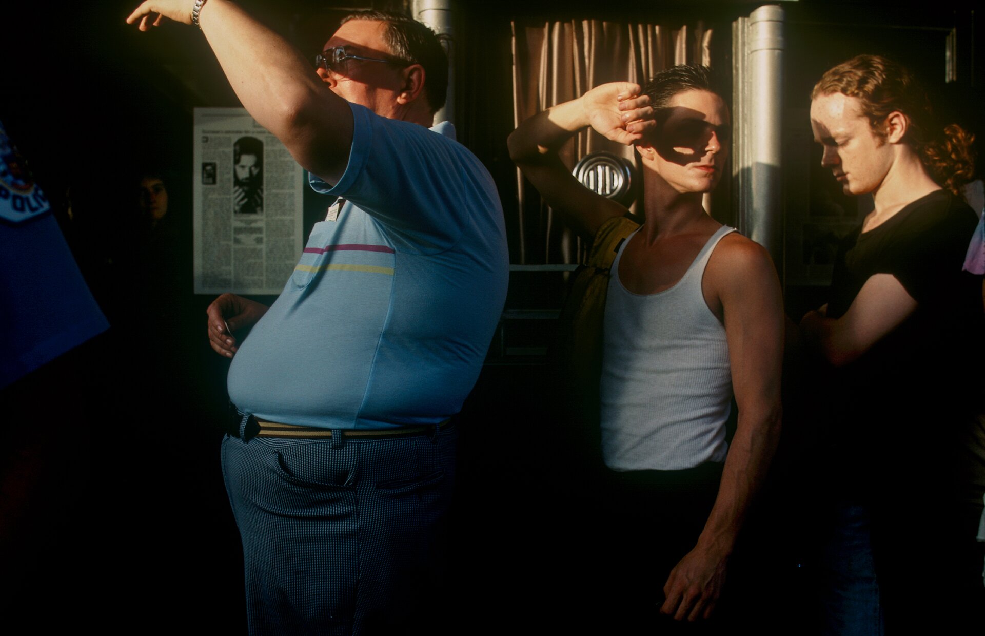 Fotografia przedstawia troje ludzi w pomieszczeniu. Mężczyźni stoją jeden za drugim, ustawieni bokiem , a pierwszy z nich to postać  z nadwagą ubrana w niebieską koszulkę z dwoma cienkimi liniami na piersi – jedną czerwoną, a drugą żółtą; niebieskie spodnie w drobne pionowe paski oraz czarno‑żółty pasek. Ma on na sobie okulary w jasnej oprawie, a na jego lewej ręce, którą zasłania się od słońca, widoczny jest srebrny zegarek. Za nim widoczna jest ściana, na której powieszono kartkę przypominającą wycinek z gazety, na której widać tekst oraz fotografię brodatego mężczyzny o długich włosach. Z lewej strony z dalszej, zacienionej, części pomieszczenia wyłania się blada twarz. Drugi z mężczyzn jest młodszy, odwraca twarz do tyłu i także jedną ręką zasłania czy przed słońcem. Jego krótkie włosy zaczesane są do tyłu, ma na sobie biały podkoszulek oraz ciemne spodnie. Jest opalony, a na jego ramieniu widać żółtą torbę. Za nim stoi trzecia postać – młody mężczyzna o długich, kręconych rudych włosach, spiętych na karku. Jego oczy skierowane są w dół, a jedną ręką obejmuje swój brzuch. Ma na sobie czarną koszulkę, a jego skóra jest blada. 