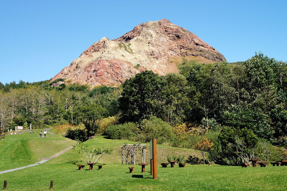 Na zdjęciu znajduje się skaliste wzniesienie. W niektórych miejscach skały mają kolor pomarańczowy. U podnóża góry teren jest zielony - rośnie trawa i drzewa. Jest ścieżka, stoją drewniane ławki. W oddali przechadzają się ludzie. Na fotografii niebo jest bezchmurne.     