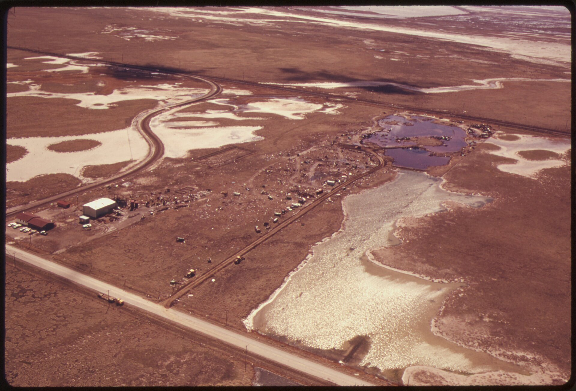 Zdjęcie wykonane z lotu ptaka przedstawia płaski teren. Ziemia ma kolor brązowoczerwony. W niektórych miejscach stoi woda. Widać trzy nieliczne budynki oraz wąską drogę. Poza tym teren jest opustoszały. 