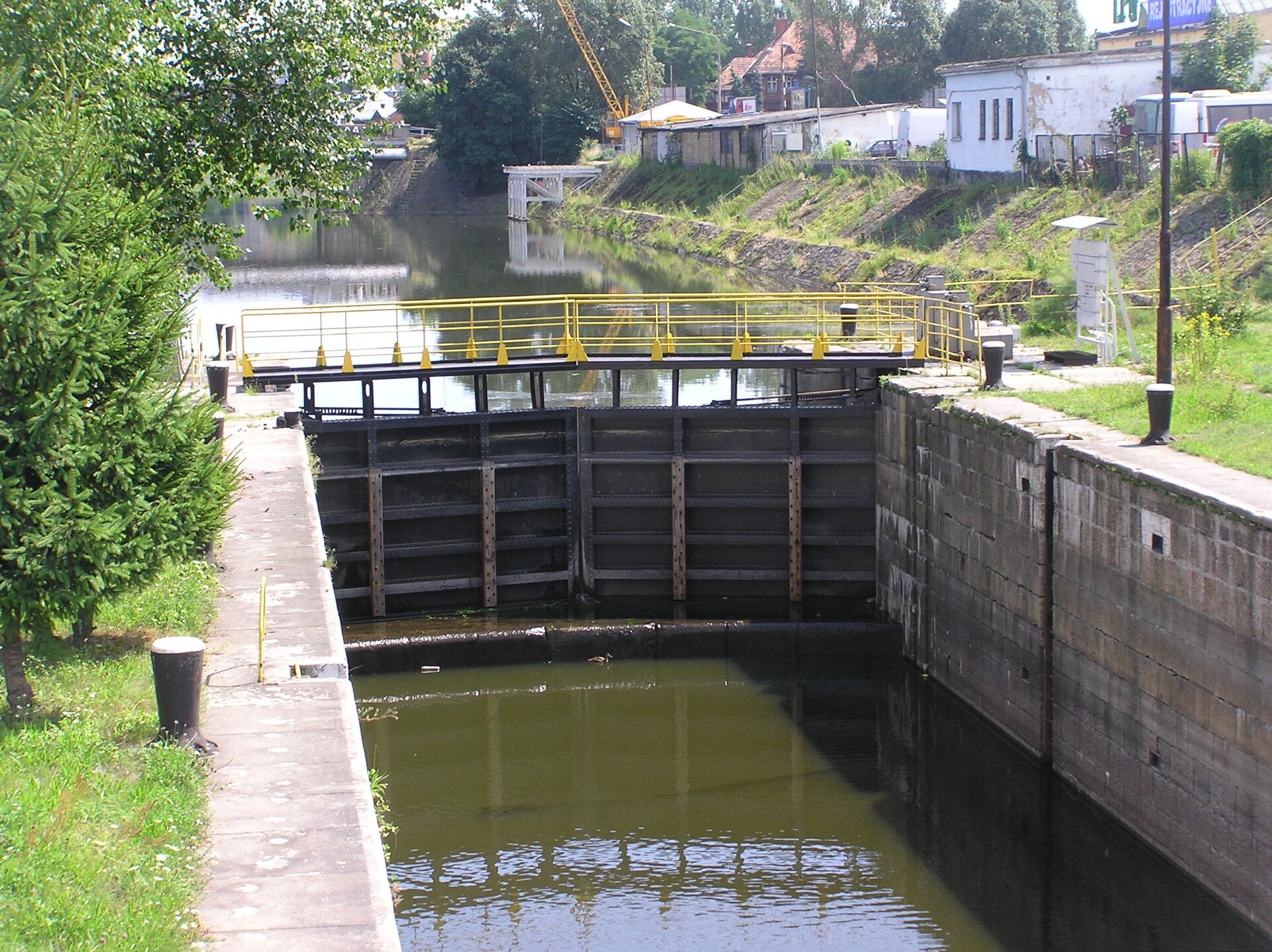 Fotografia prezentuje śluzę na kanale łączącym dwa zbiorniki o różnym poziomie wody. Śluza skład się z wrót, które zamykają się na kanale, zatrzymując przepływ wody.