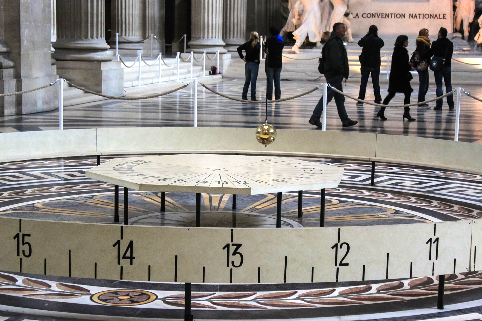 Fotografia przedstawia główną część wahadła Foucaulta, którą stanowi wielka metalowa kula zwisająca na długiej lince. Na podłodze widać obramowanie ze skalą. Na drugim planie postacie ludzkie i ściany budynku.